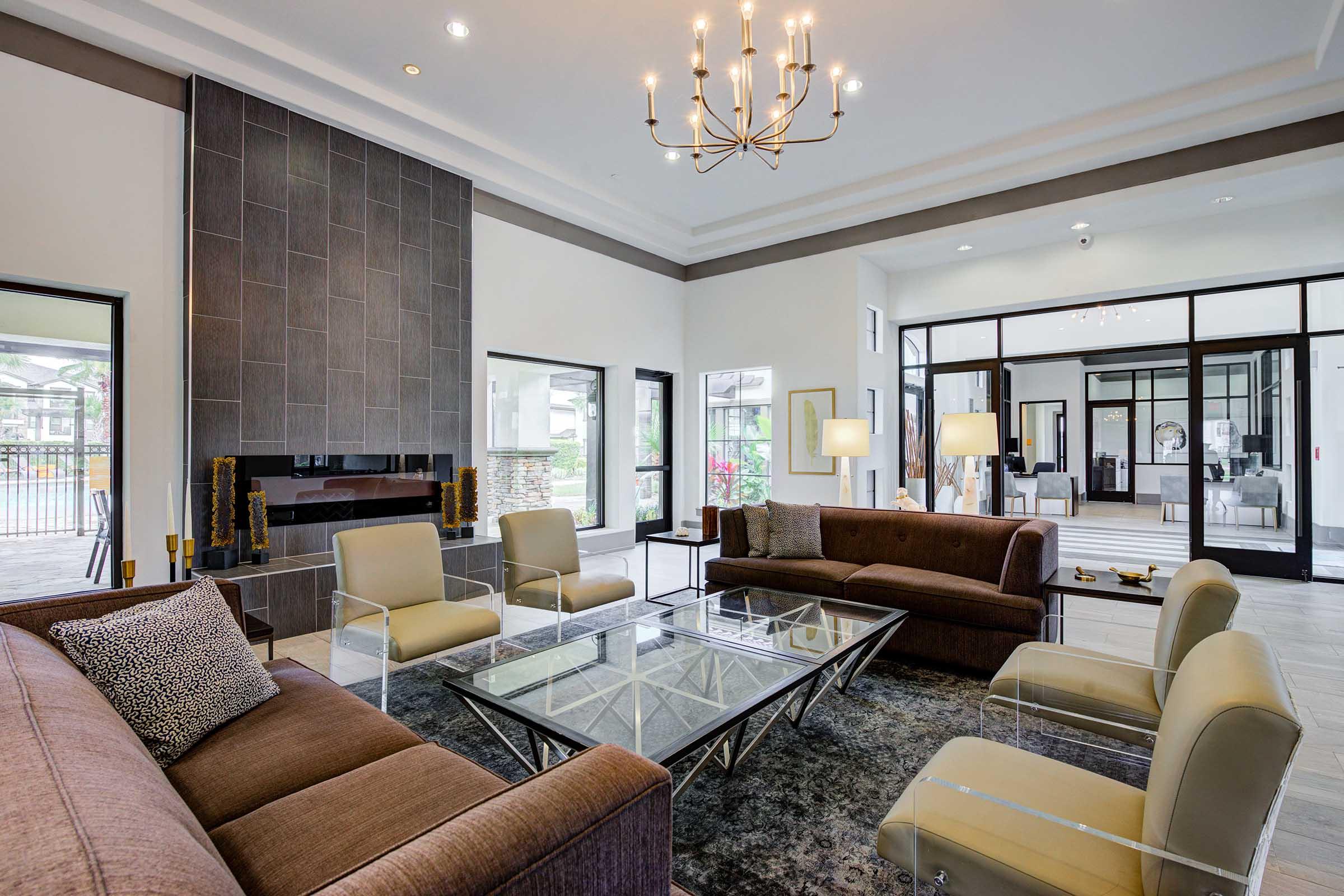 A modern living room featuring a stylish design, with a large glass coffee table at the center, two brown sofas, and light-colored armchairs. The room has a sleek stone fireplace and large windows allowing ample natural light. Elegant lighting fixtures hang from the ceiling, enhancing the contemporary ambiance.