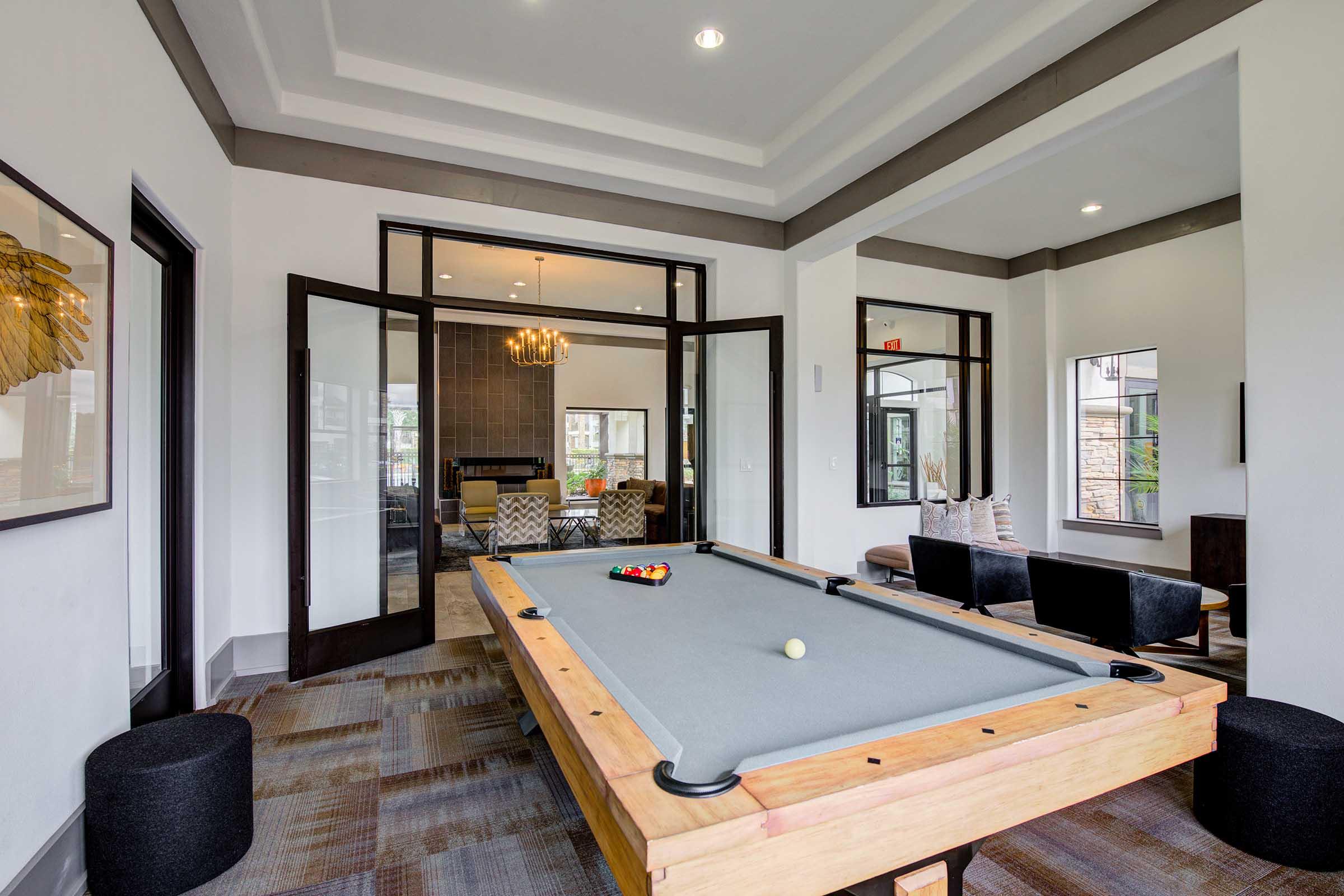 A well-lit room featuring a pool table in the foreground, with a gray felt surface and a single white ball. In the background, there are large windows and stylish seating arrangements. The modern decor includes a chandelier and wall art, creating a welcoming atmosphere for leisure activities.