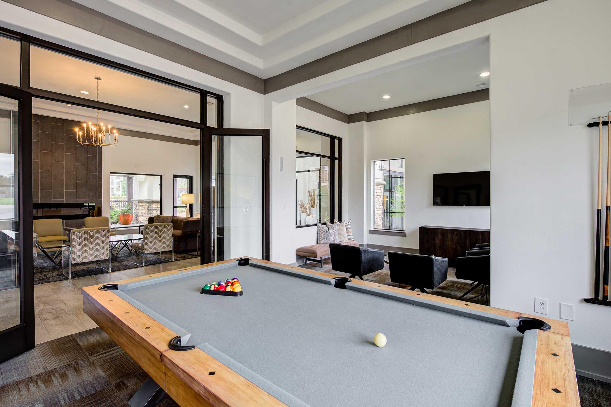 A modern recreation room featuring a pool table in the foreground. The room has large windows, stylish furniture, and a television on the wall. There are decorative light fixtures and a comfortable seating area visible in the background. The decor is contemporary and inviting.