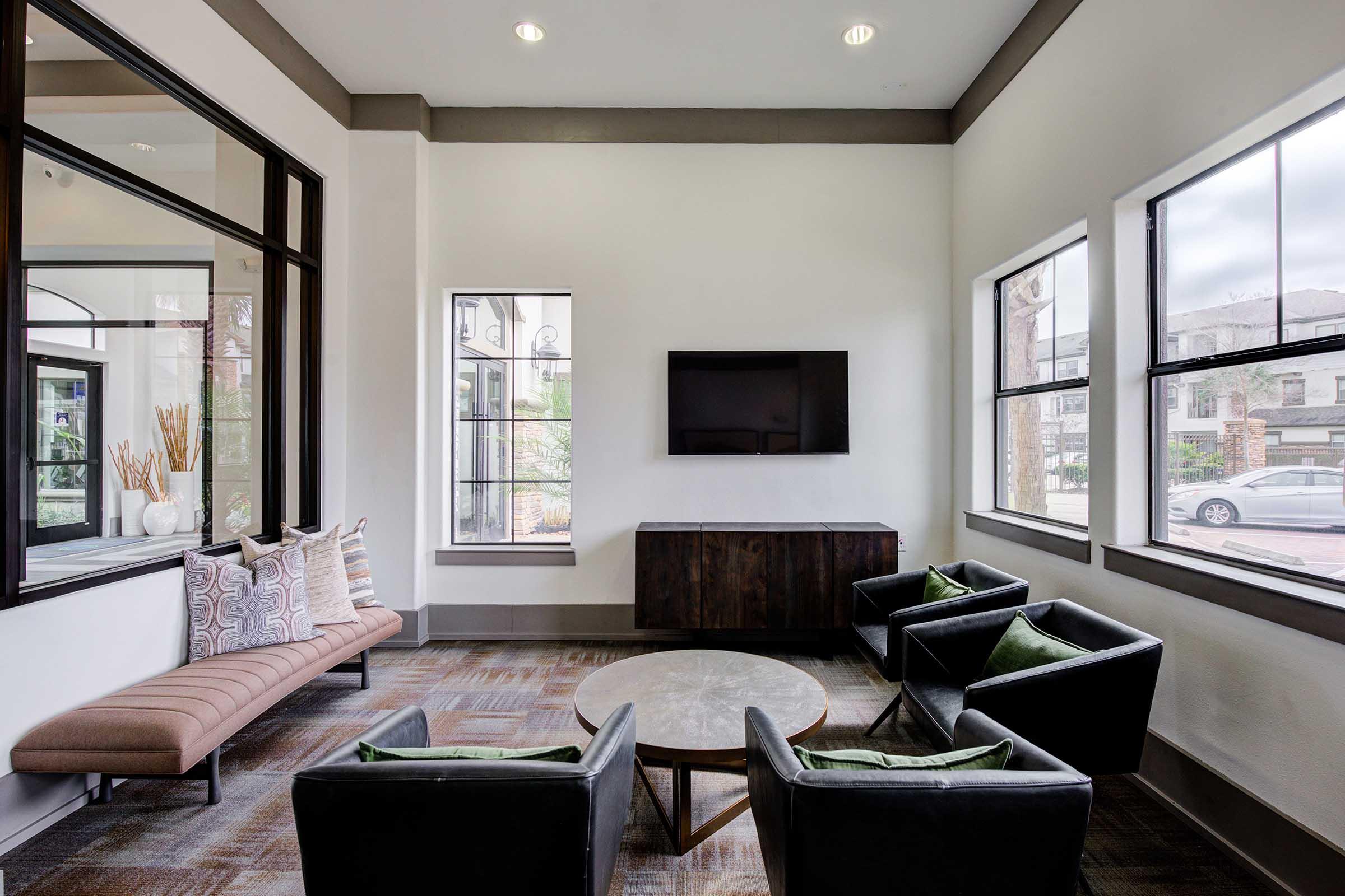 A modern lounge area featuring a comfortable bench with decorative pillows, a circular coffee table, and four black armchairs with green cushions. There is a flat-screen TV mounted on the wall, and large windows allow natural light to brighten the space, creating a welcoming atmosphere.