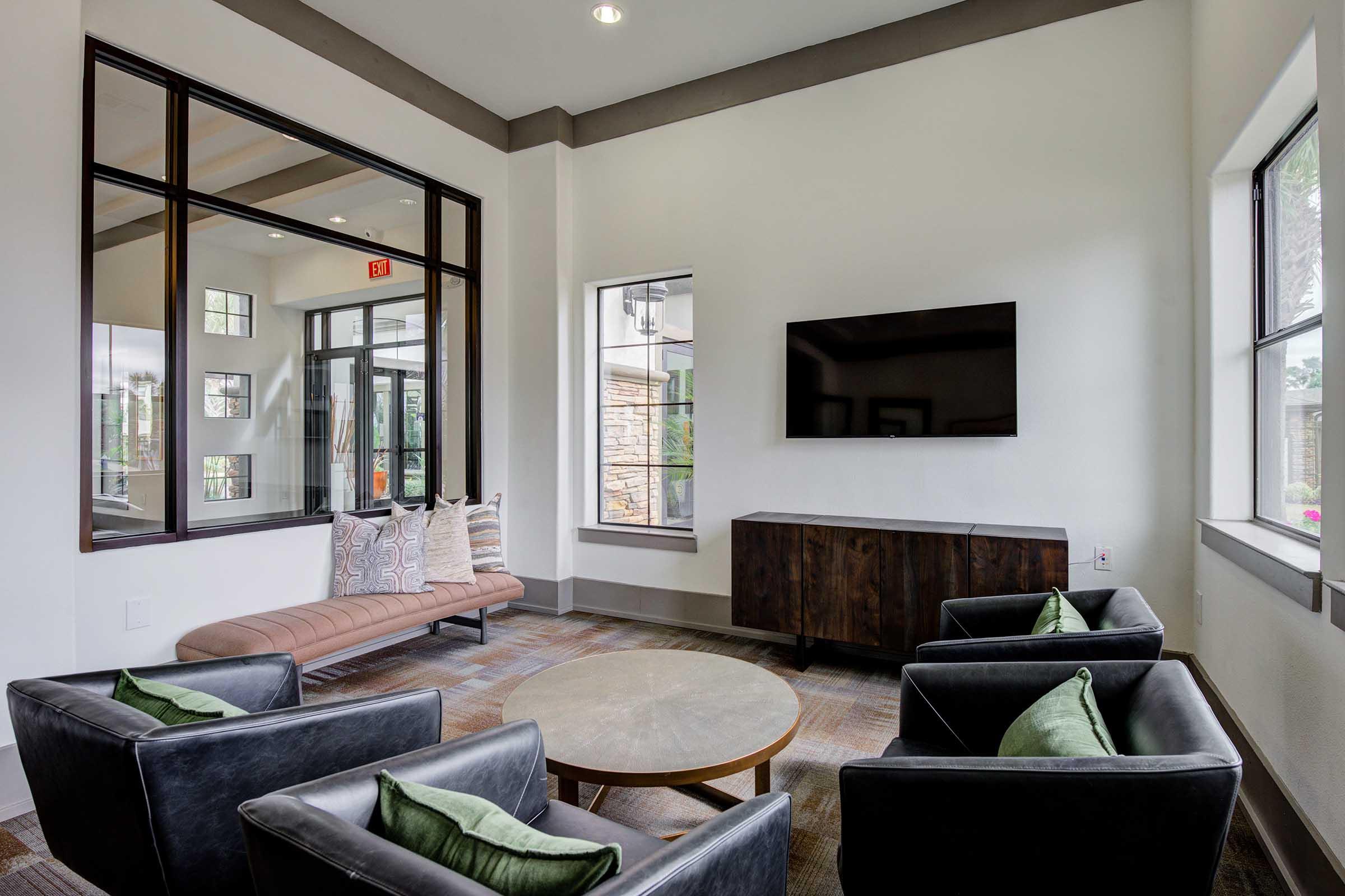 A modern lounge area featuring a light-colored wall, large windows, and a flat-screen TV mounted on a wooden cabinet. There are four black armchairs with green cushions arranged around a round coffee table, and a long, cushioned bench against one wall, creating a comfortable and inviting space.