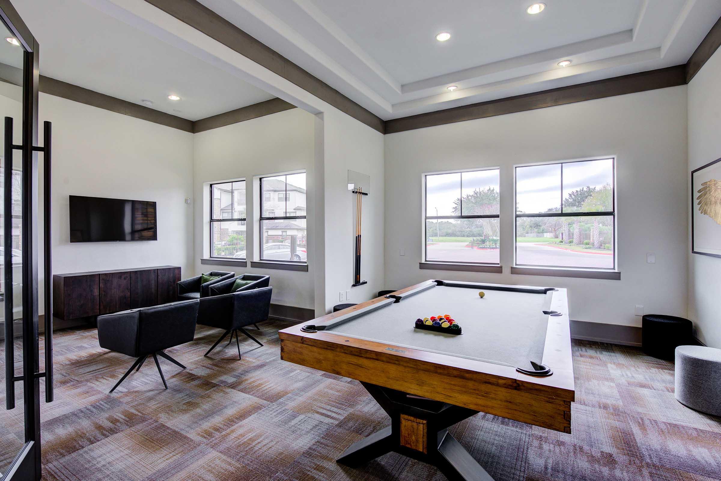 A stylish recreational room featuring a billiards table with colorful balls set on it. Surrounding the table are modern black armchairs and a dark wood cabinet. Large windows let in natural light, illuminating the space. A flat-screen TV is mounted on the wall, enhancing the room's entertainment appeal.