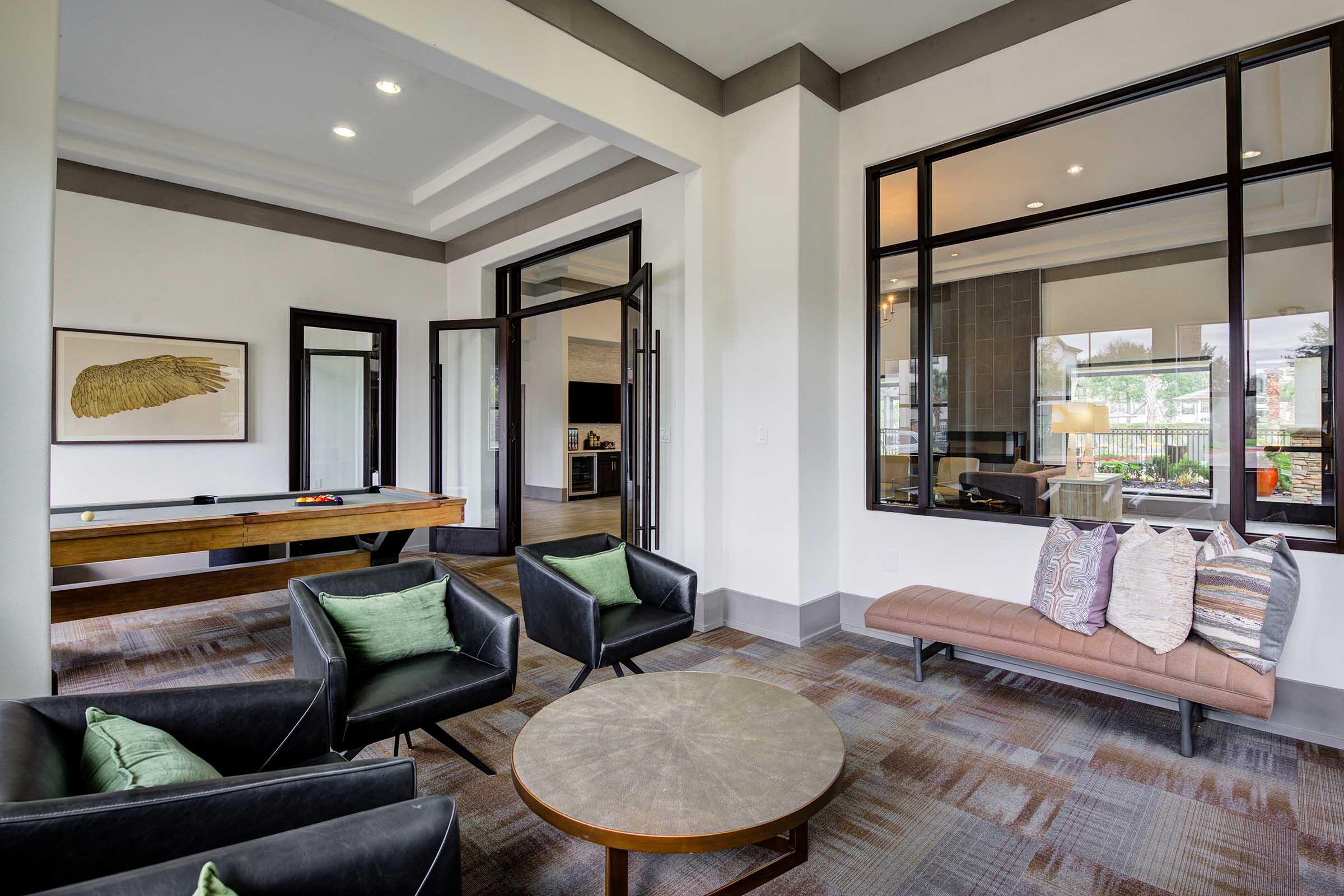 A modern lounge area featuring a pool table, plush black chairs with green cushions, and a round coffee table. Large windows allow natural light to fill the space, which has a contemporary design with neutral tones and stylish decor.