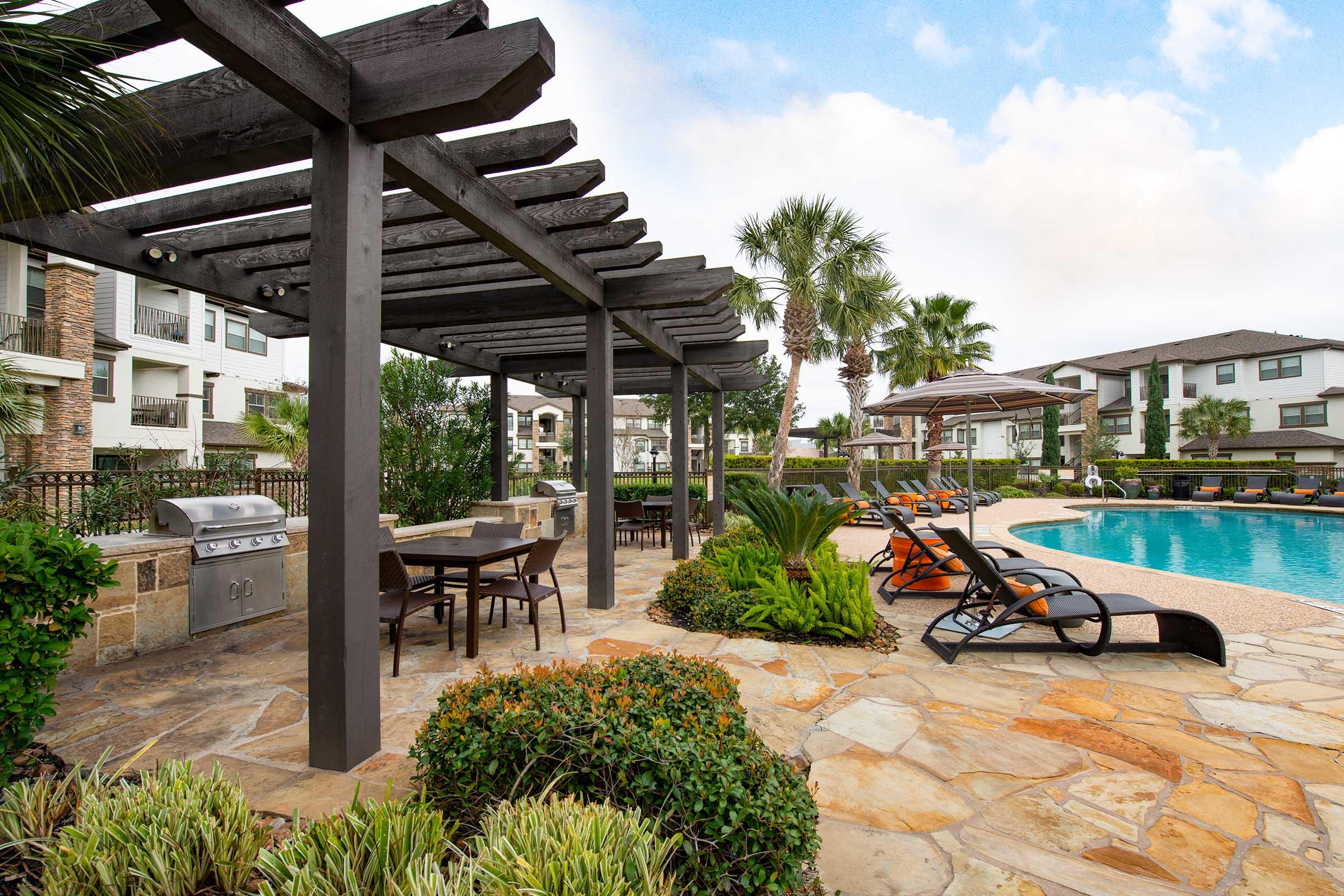 A serene outdoor pool area featuring a large, inviting swimming pool surrounded by lounge chairs. There are shaded pergolas with outdoor dining tables and a grill, set among lush green landscaping and palm trees. Modern apartment buildings are visible in the background under a partly cloudy sky.