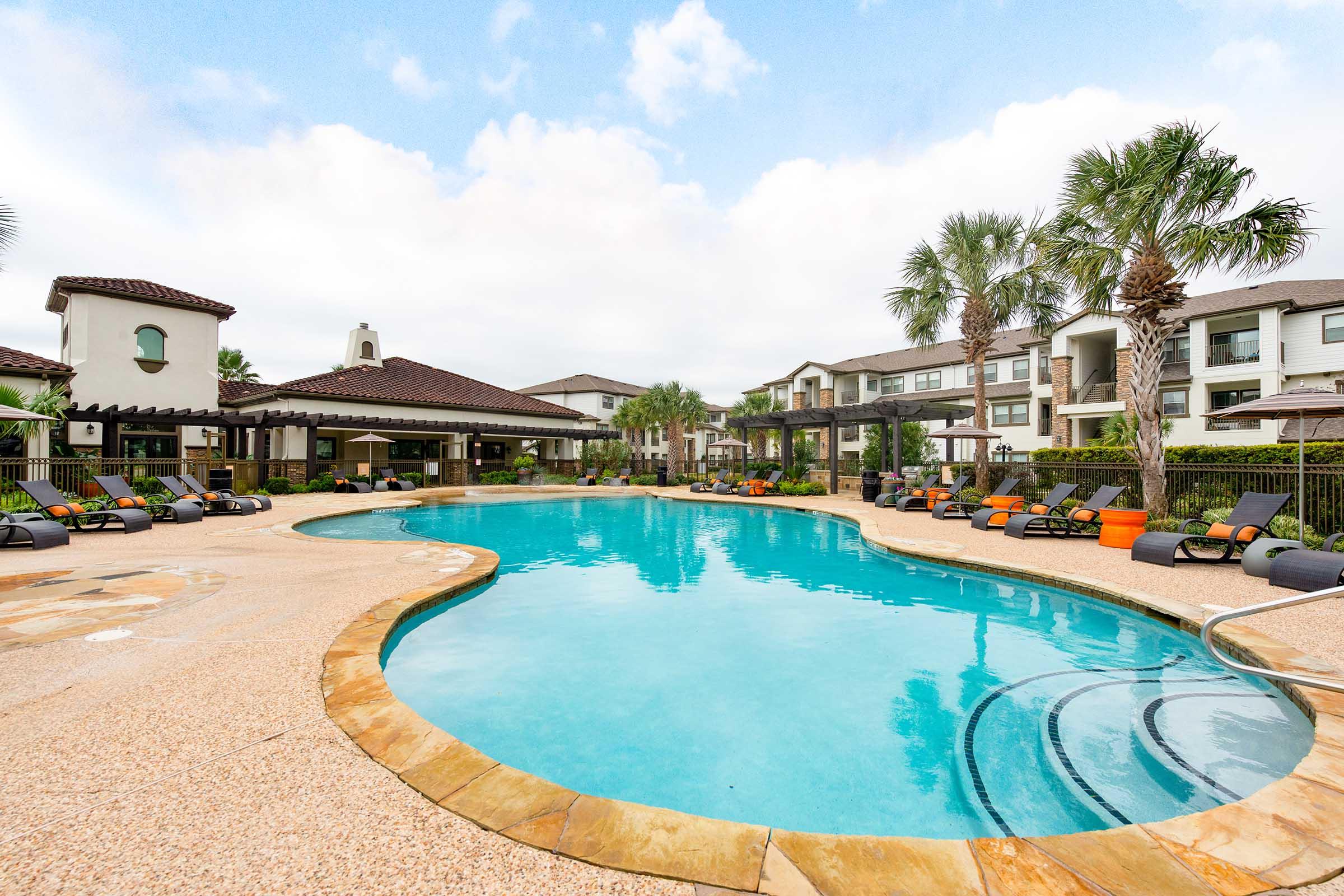 A spacious outdoor pool surrounded by lounge chairs and palm trees, with modern apartment buildings in the background. The sky is partly cloudy, creating a serene and inviting atmosphere for relaxation and leisure.