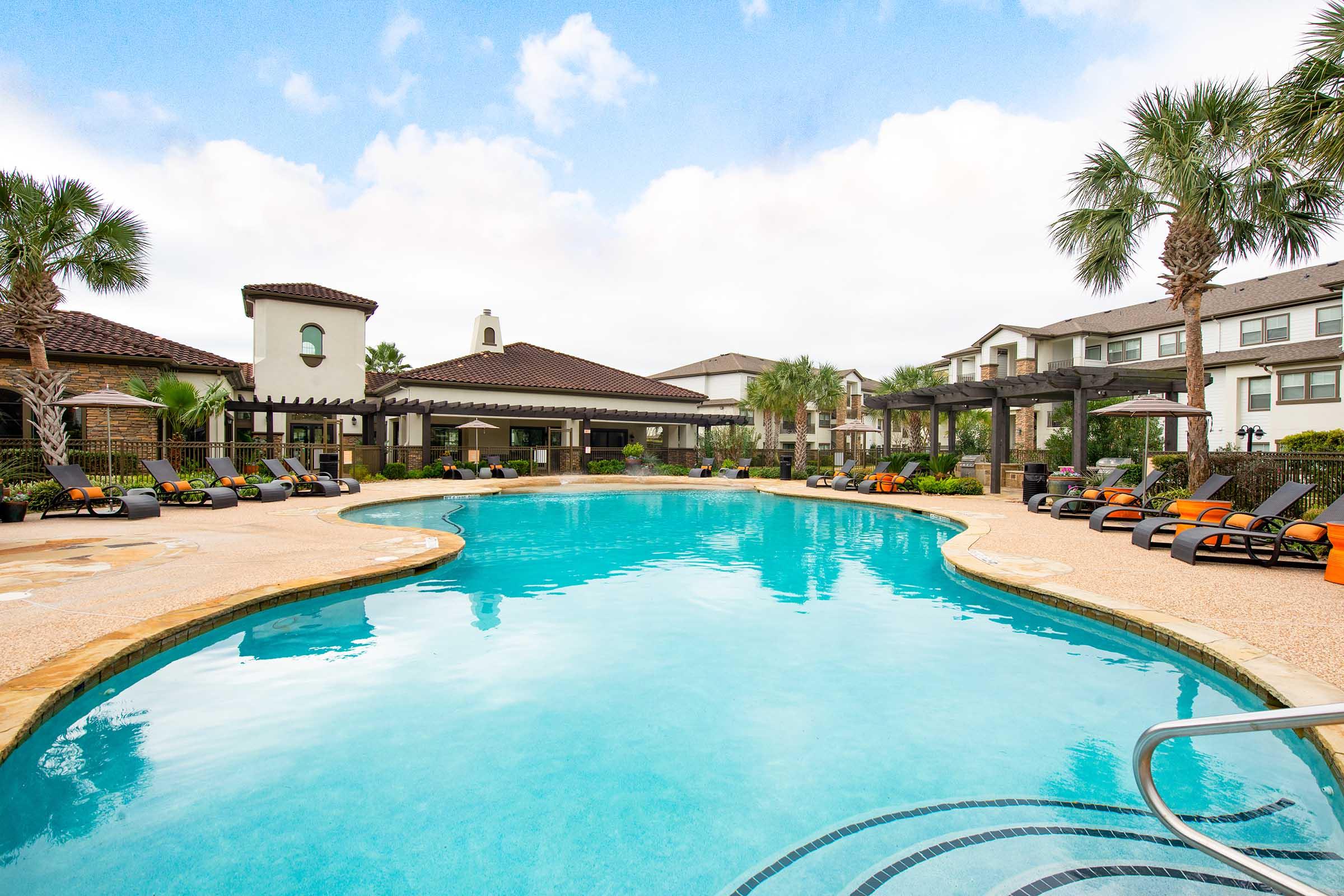 A spacious swimming pool surrounded by lounge chairs, palm trees, and well-manicured landscaping. The area features modern architecture with a building in the background, adding to the resort-like atmosphere under a partly cloudy sky.