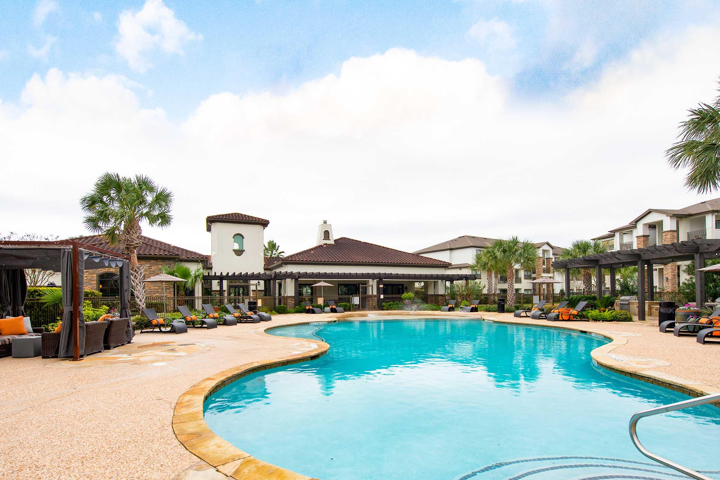 A large outdoor swimming pool surrounded by lounge chairs and cabanas, with palm trees and landscaped gardens. In the background, there are modern residential buildings with a Mediterranean architectural style, and a cloudy sky above. The scene conveys a relaxing and inviting atmosphere.