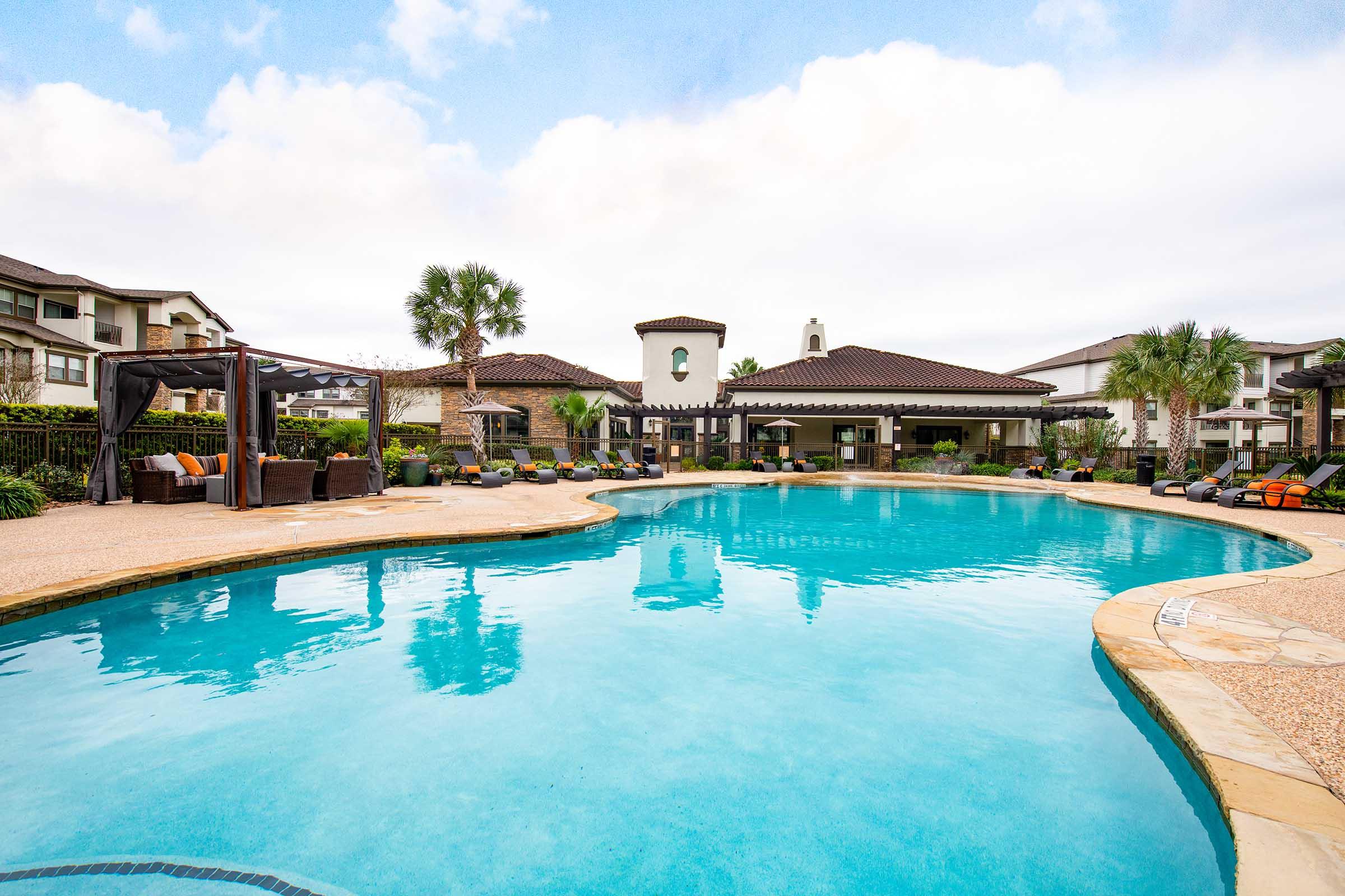 A tranquil outdoor swimming pool surrounded by palm trees and lounge chairs, featuring a stylish gazebo area with seating. The pool area is complemented by well-maintained landscaping and modern buildings in the background, all set under a partly cloudy sky.