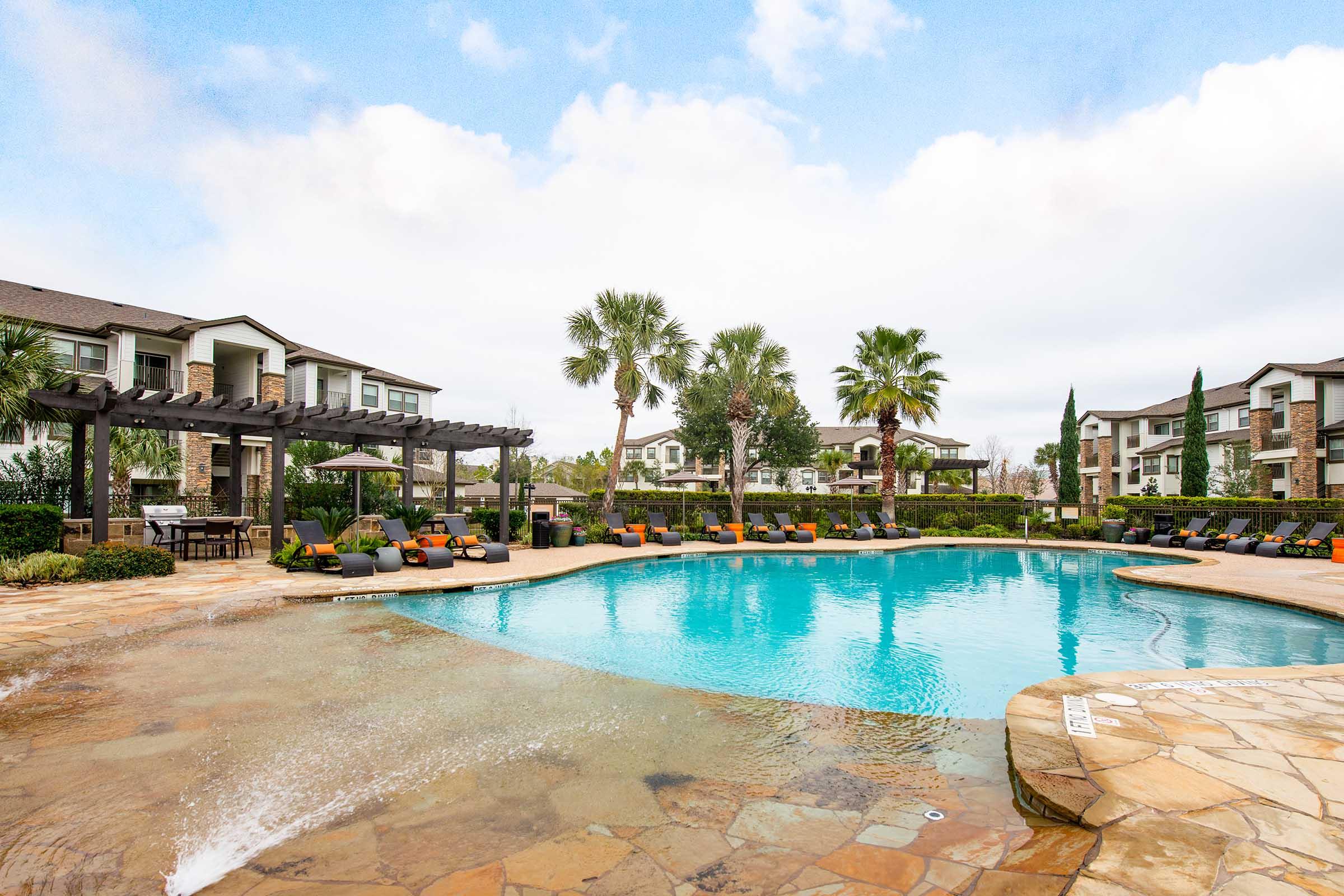 A spacious outdoor swimming pool surrounded by lounge chairs and palm trees, with modern apartment buildings in the background. The pool area features landscaping and is under a partly cloudy sky, creating a relaxing atmosphere.