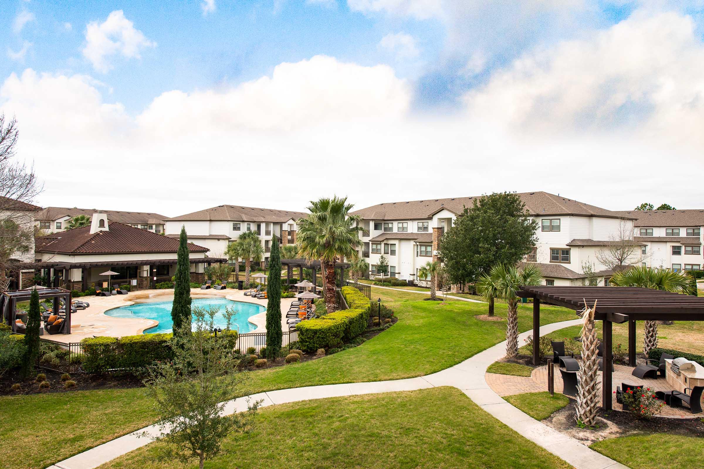 A scenic view of a landscaped apartment community featuring a large swimming pool, surrounded by palm trees and lounge chairs. There are shaded pergolas and walking paths, with well-maintained green lawns and a clear blue sky in the background.