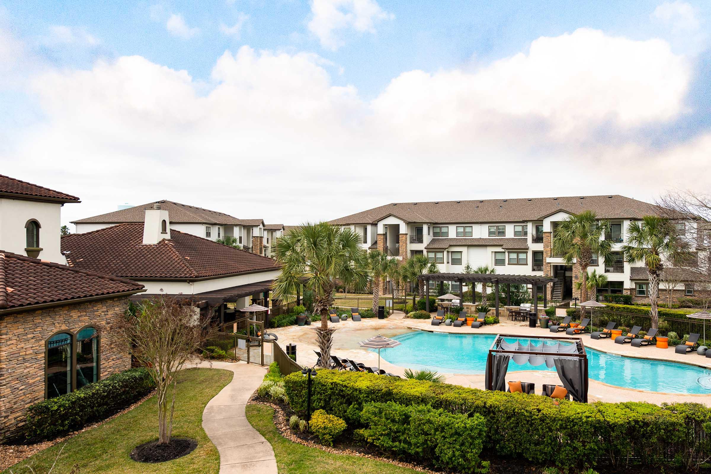 A view of a modern apartment community featuring a pool surrounded by lounge chairs and palm trees. The landscape includes well-maintained greenery and a path leading to the pool area, with buildings in the background under a cloudy sky.