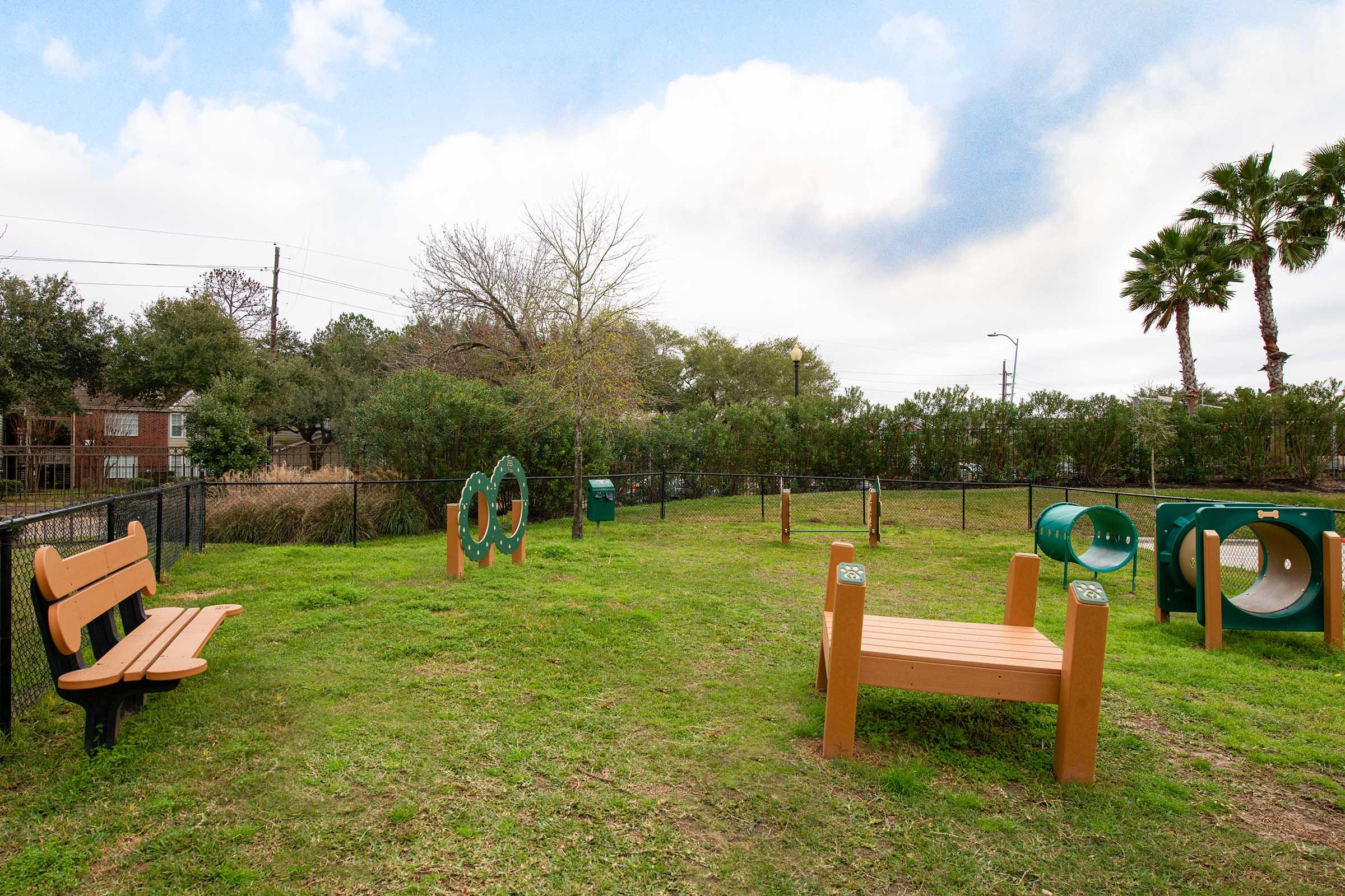 A grassy play area featuring a couple of wooden benches and green playground structures, including tunnels and hoops. The background includes shrubs and palm trees, with a fence surrounding the space. The sky is partly cloudy, creating a bright outdoor atmosphere.