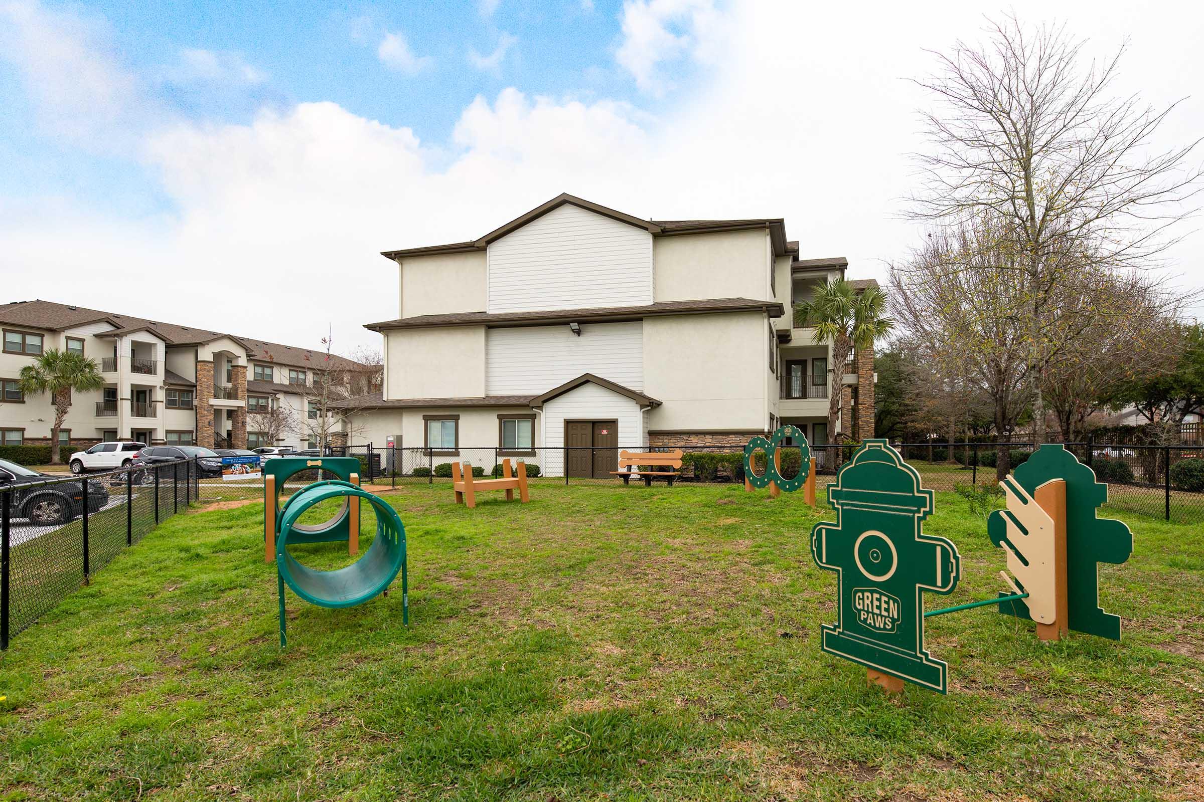 A grassy area in front of an apartment building featuring a small dog park. The park includes a green fire hydrant, a tube tunnel, and various playful dog-themed structures. There are benches and a fence surrounding the space, with cars parked nearby and trees in the background under a cloudy sky.