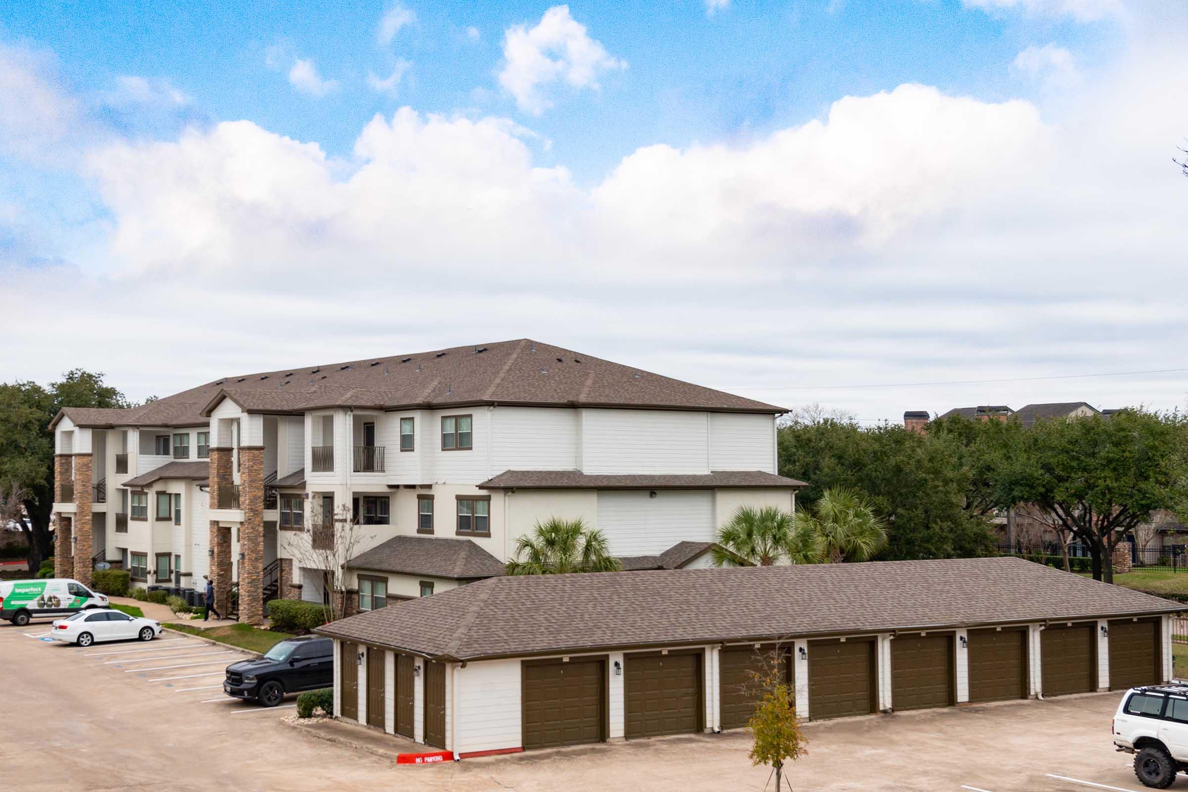 A multi-story residential building with a mix of stone and siding exterior. The building is surrounded by trees and features several parking spaces with vehicles. In the foreground, there are storage garages with green doors. The sky is partly cloudy.