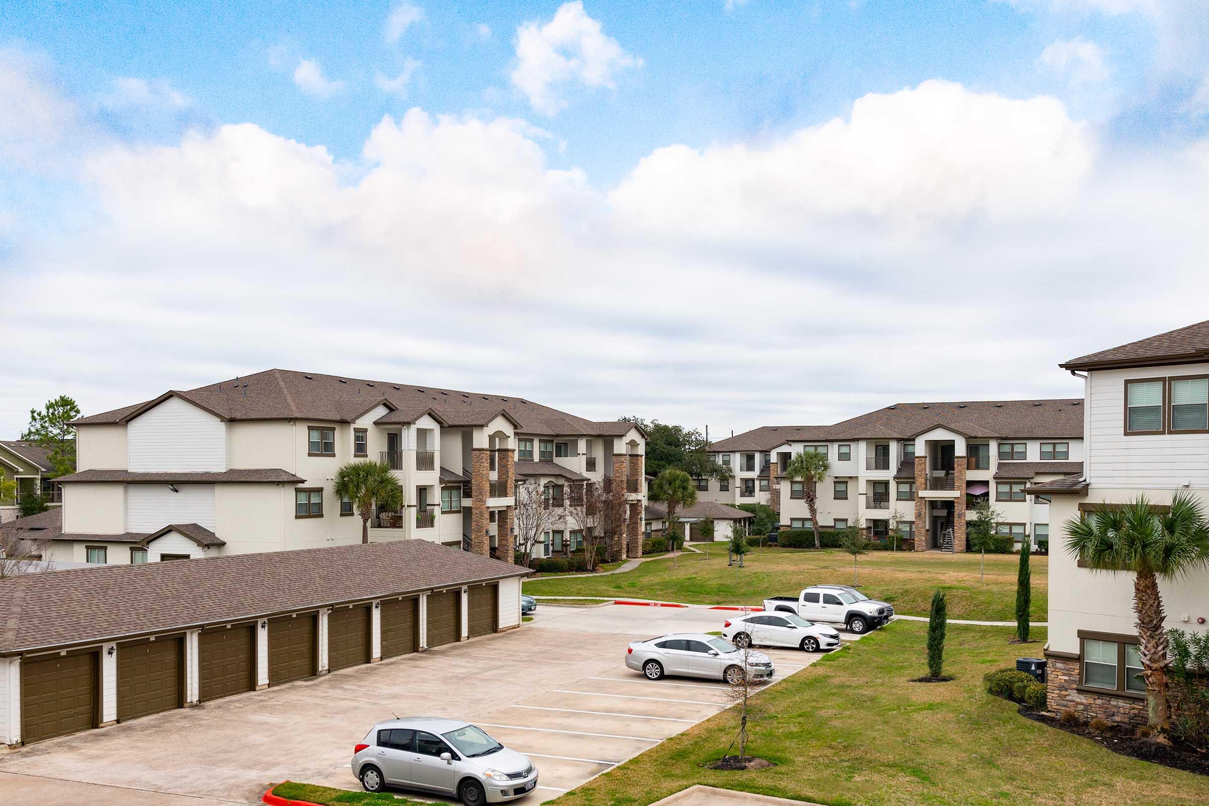 View of a residential apartment community featuring multiple multi-story buildings. The scene includes well-maintained lawns, parked cars in a lot, and palm trees. The sky is partly cloudy, creating a serene atmosphere.