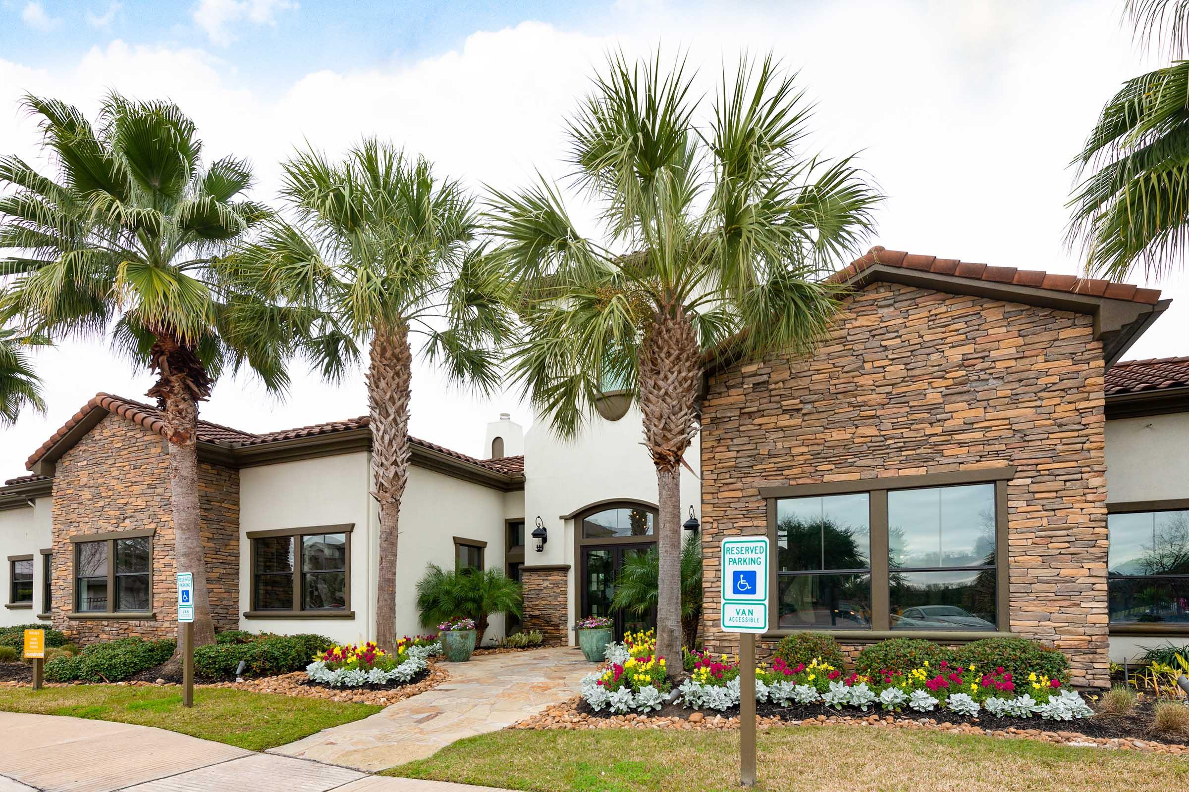 A modern building with a stone exterior, featuring large windows, surrounded by palm trees and colorful flower beds. A sign indicates accessible parking. The sky is partly cloudy, adding to the inviting atmosphere of the property.