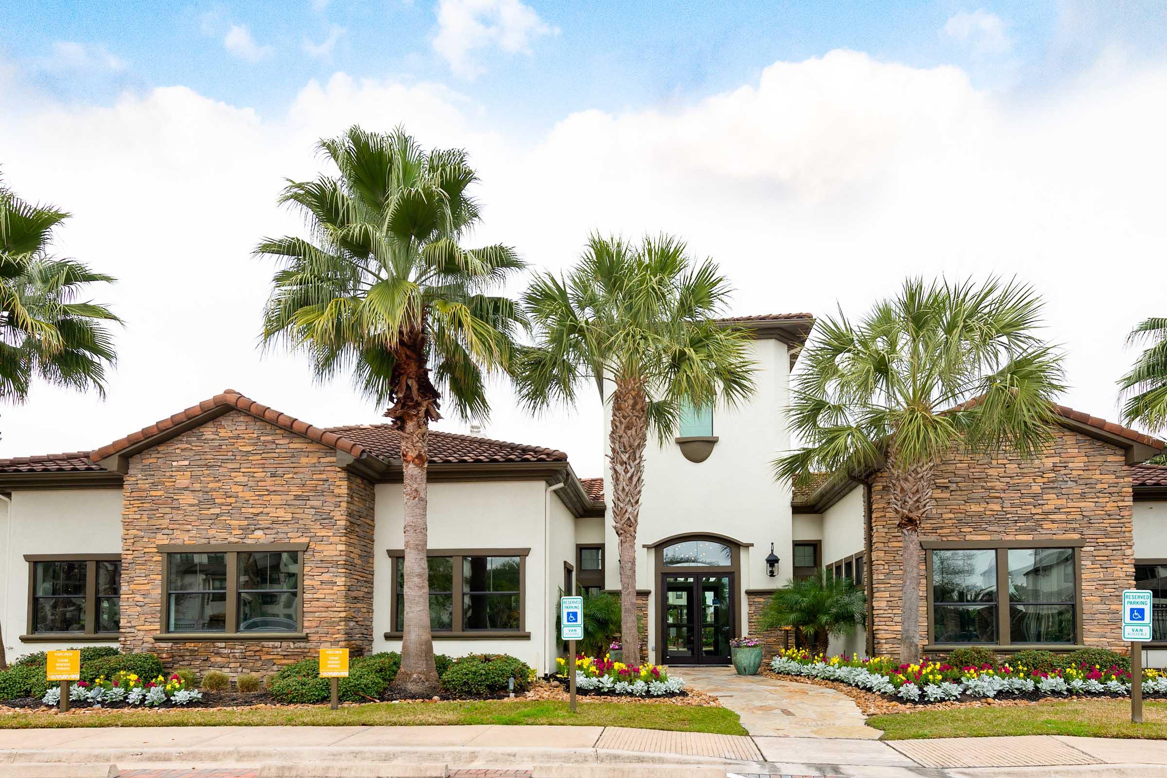 A modern building with a combination of stone and smooth white walls, featuring large windows. Palm trees and colorful flower beds surround the entrance, which has a decorative pathway leading up to double doors. The sky is partly cloudy, enhancing the inviting atmosphere of the property.