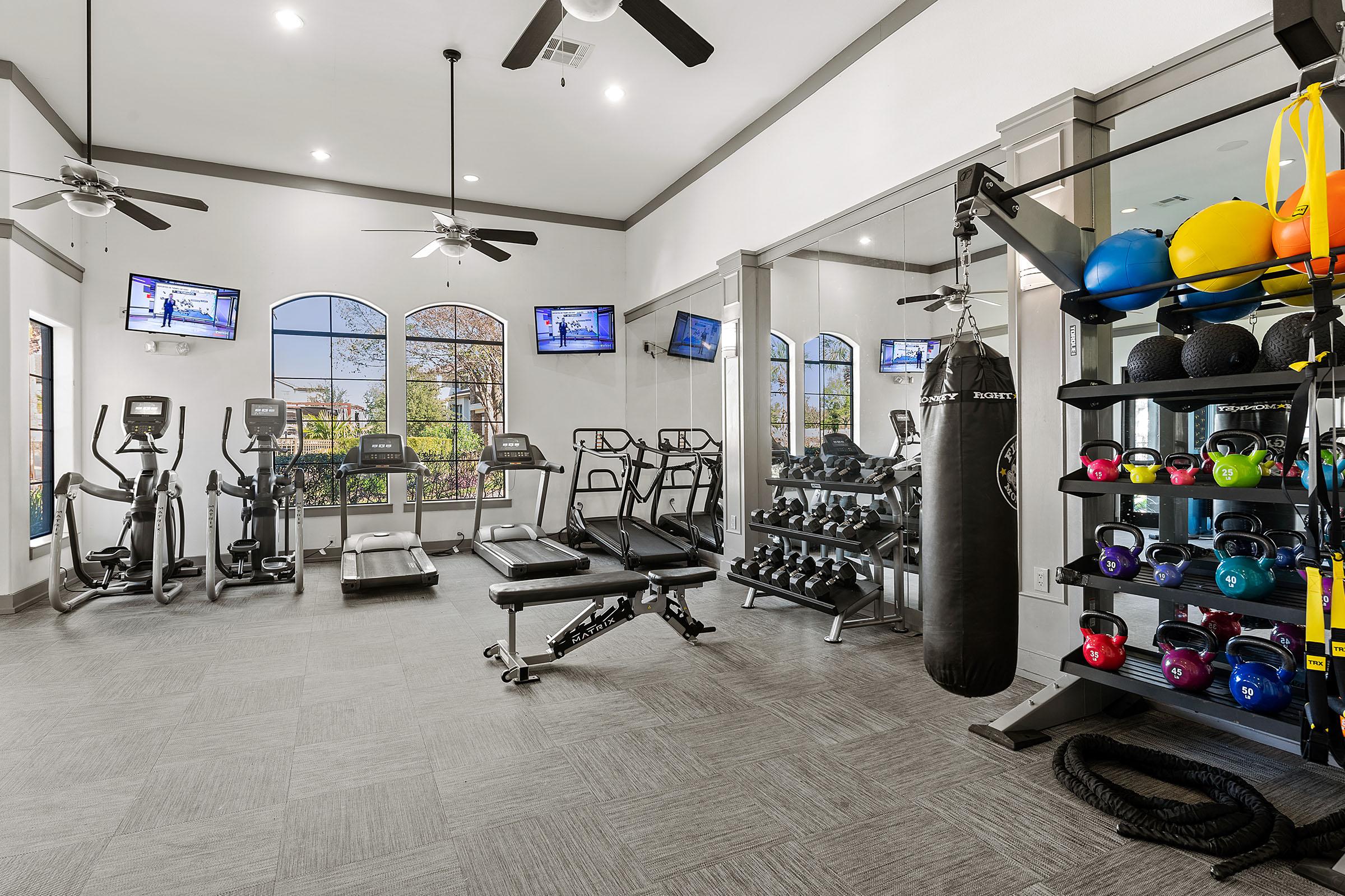 A modern gym featuring cardio equipment like treadmills and stationary bikes, a set of dumbbells, a weight bench, and a punching bag. Large windows provide natural light, and multiple televisions are mounted on the walls for entertainment. The space is well-lit and designed for fitness activities.