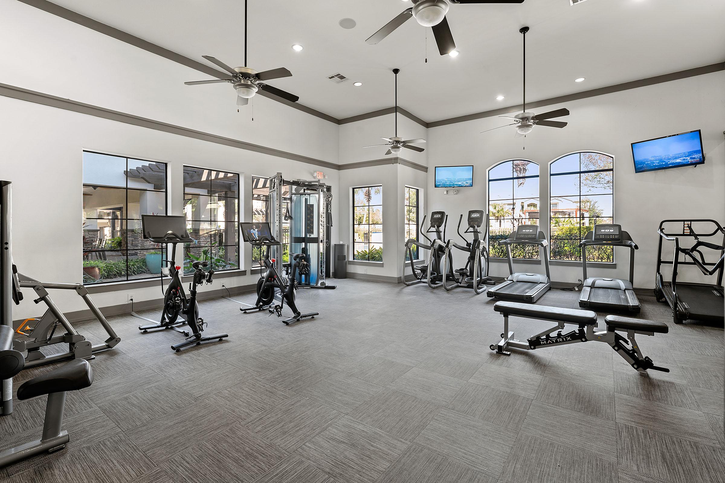 A well-lit fitness room featuring various exercise equipment, including treadmills, stationary bikes, weight benches, and a multi-gym station. Large windows provide natural light and views of outdoor greenery, with ceiling fans and multiple wall-mounted screens for viewing.