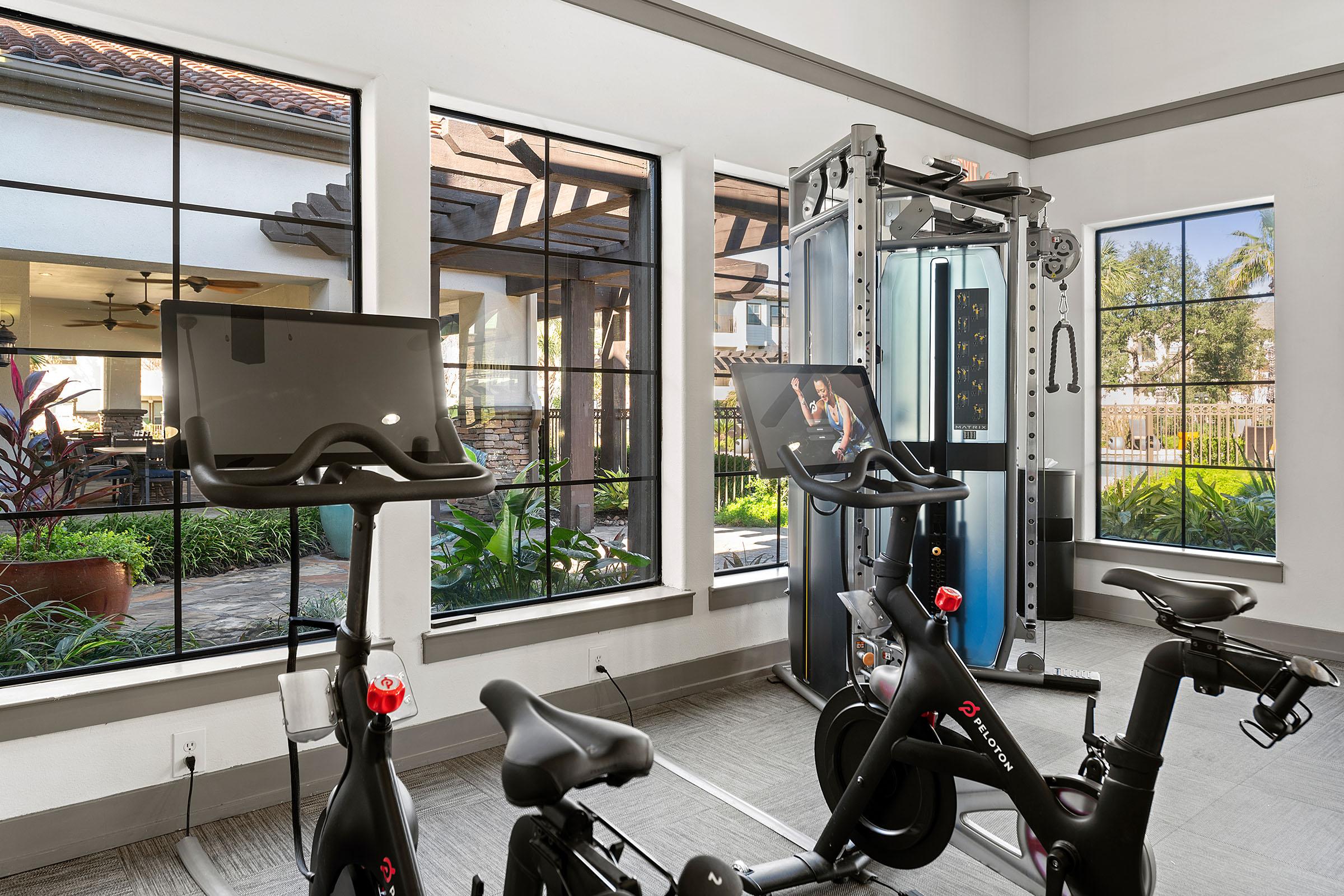 A modern gym space featuring exercise bikes with large screens, a cable machine, and large windows allowing natural light to fill the room. The outdoor area includes lush greenery visible through the windows, enhancing the overall ambiance of the fitness environment.