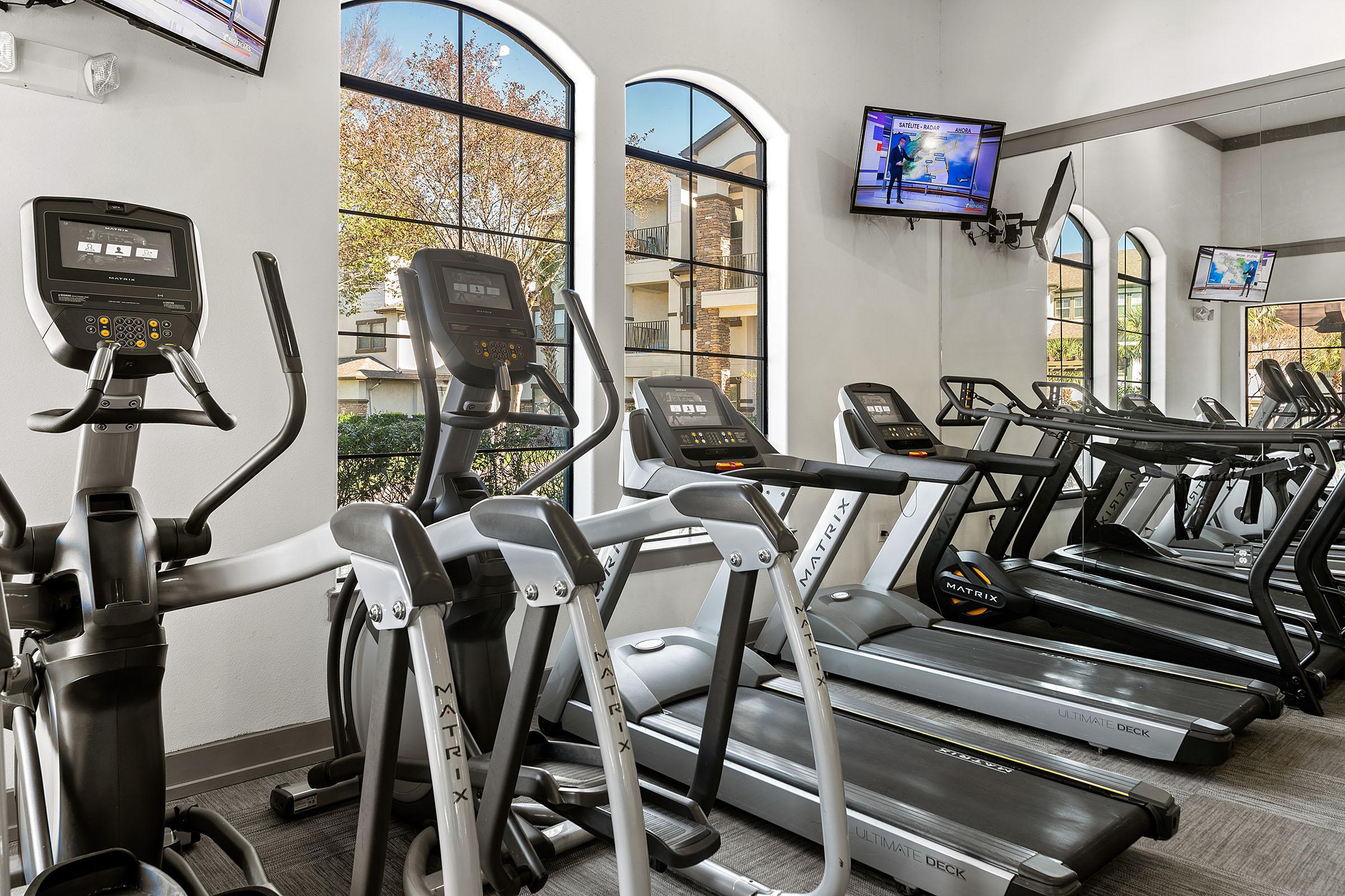 A modern fitness center featuring several elliptical machines and treadmills lined up against large windows. Two TVs are mounted on the wall, displaying news or workout content, with natural light streaming in from the windows. The space is well-lit and inviting for exercise.