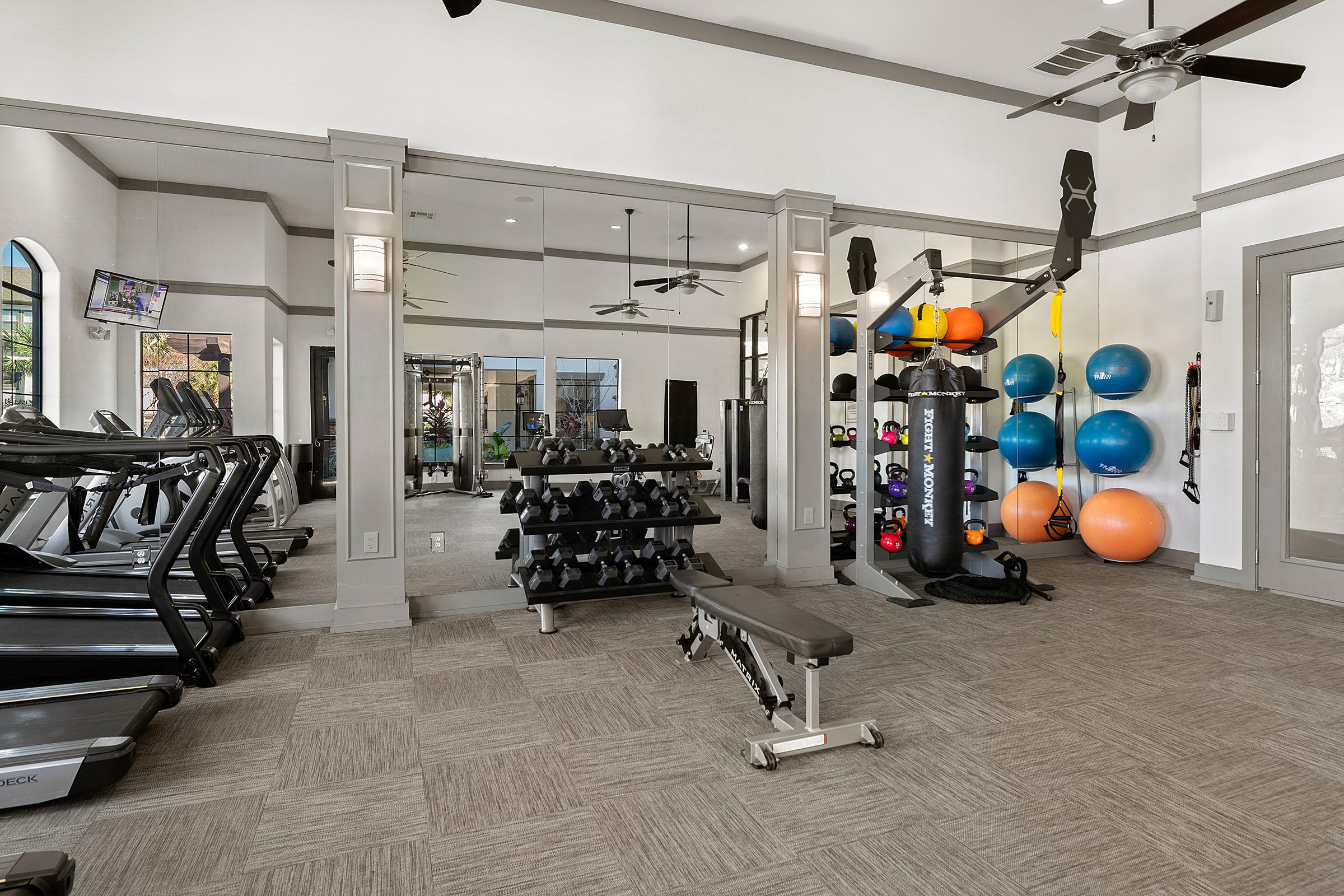 A modern fitness center featuring treadmills, a weight bench, a rack of free weights, and exercise balls. The space is well-lit with large mirrors and windows, creating an inviting atmosphere for workouts.