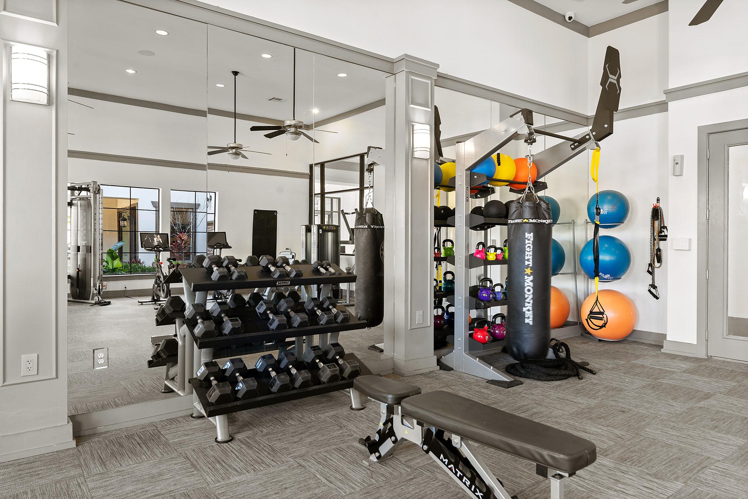 A well-lit modern gym featuring a variety of fitness equipment. There's a weight bench, a set of dumbbells, a punching bag, and a multi-station weight machine. Colorful exercise balls and a cable machine are also visible, with large mirrors reflecting the space. The decor is sleek and contemporary.