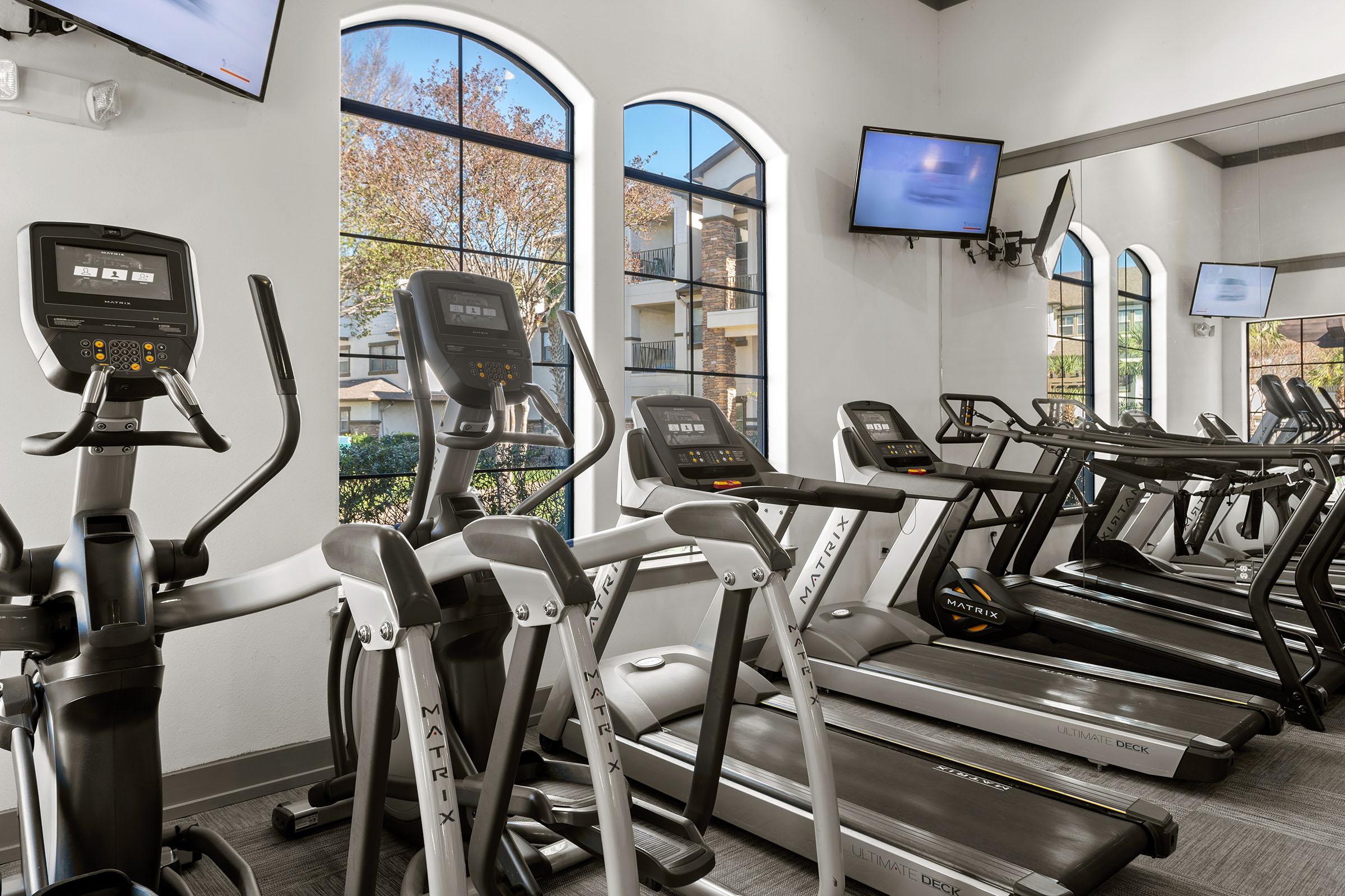 A modern gym interior featuring several Matrix treadmills and elliptical machines facing large arched windows. Natural light streams in, illuminating the space, which includes two wall-mounted televisions and a view of greenery outside. The atmosphere is clean and inviting, ideal for workouts.