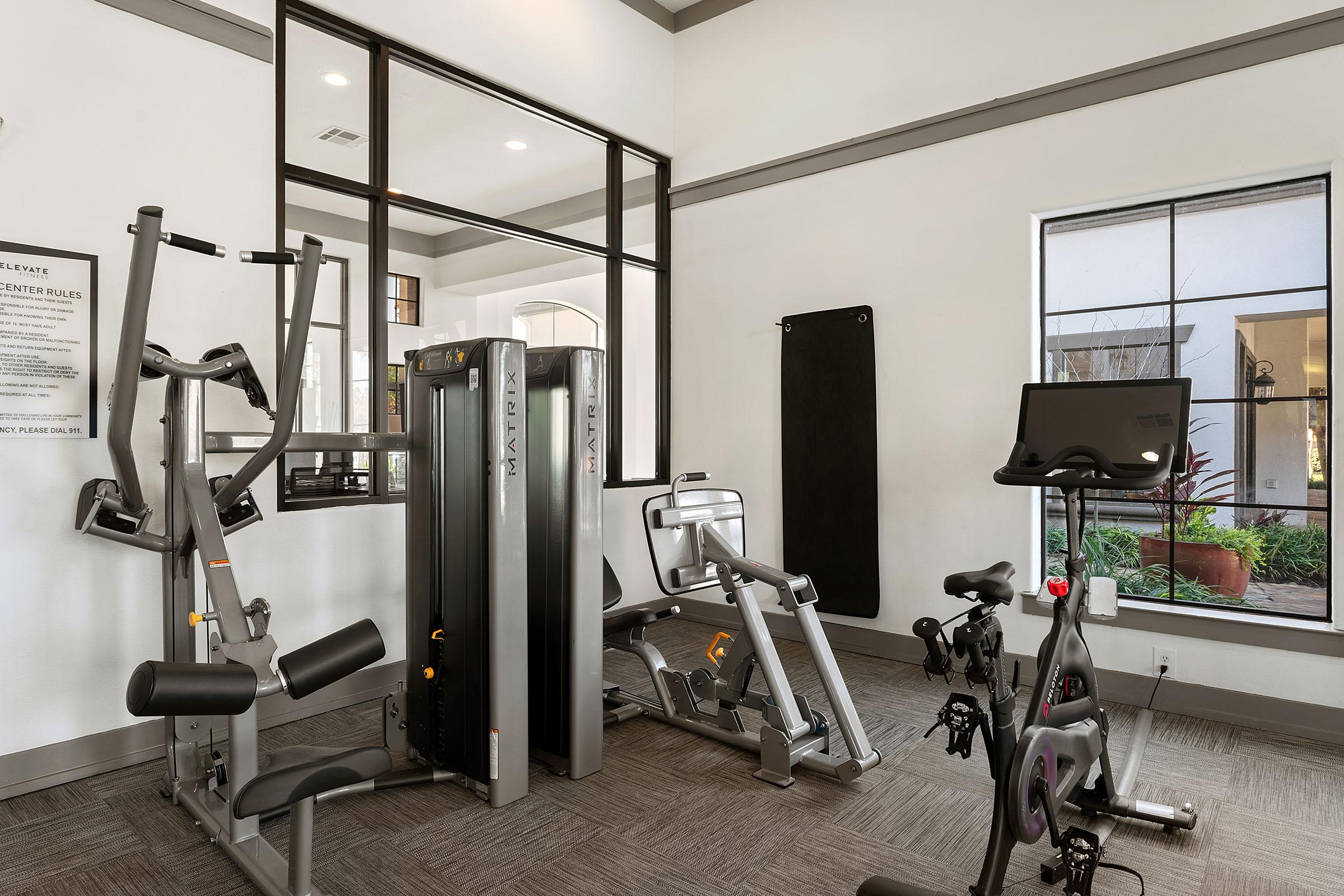 A modern gym interior featuring various exercise equipment, including a multi-gym machine, a stationary bike, and a wall-mounted mirror. Natural light enters through large windows, highlighting the clean and spacious environment designed for fitness activities.