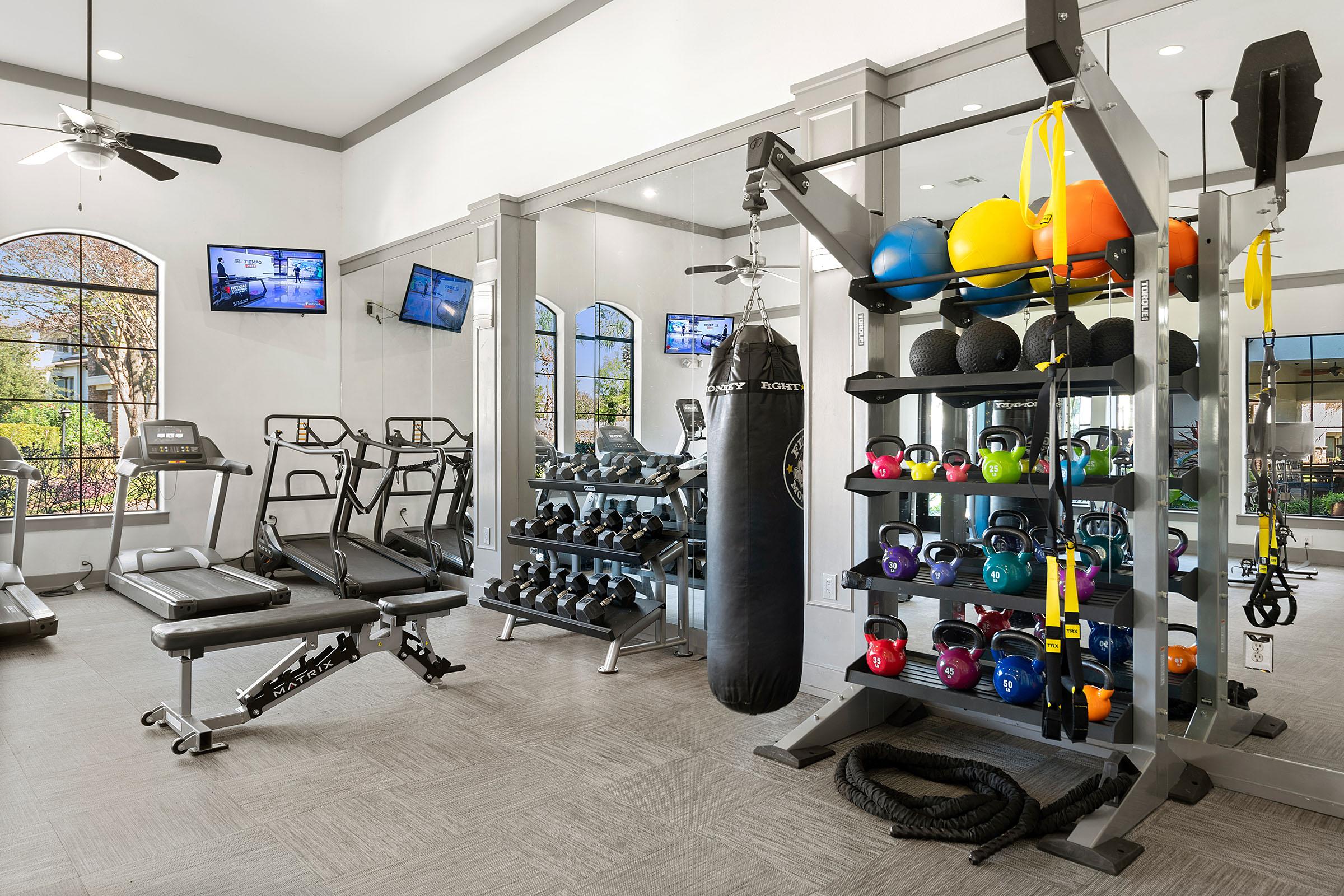 A modern gym interior featuring workout equipment, including a weight bench, dumbbell rack, exercise machines, a punching bag, and a cable machine with various colored kettlebells. Large windows let in natural light, and two wall-mounted screens are visible in the background.