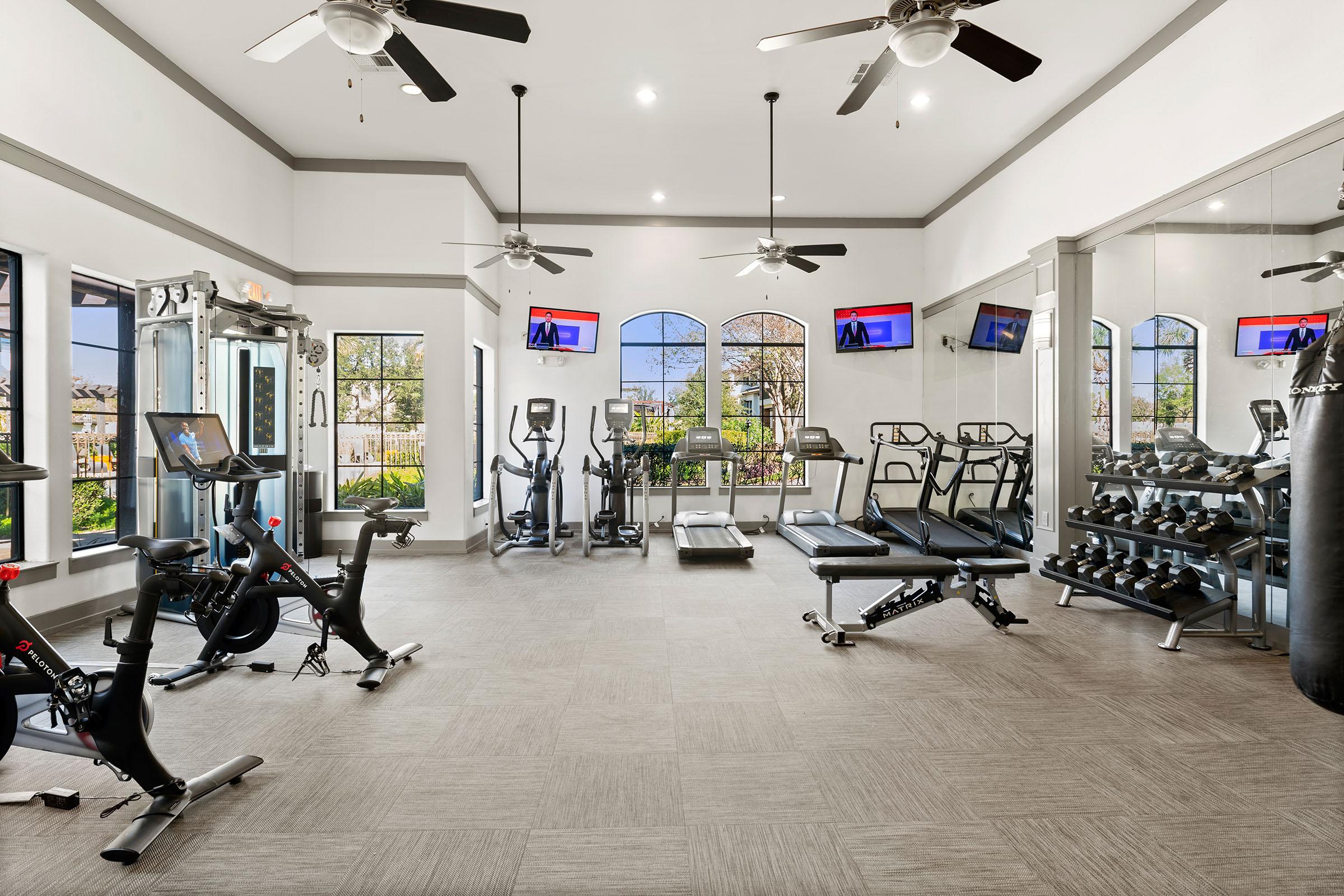 A modern gym interior featuring exercise equipment such as treadmills, stationary bikes, and weight machines. The space has large windows allowing natural light, ceiling fans for ventilation, and two television screens mounted on the walls. The flooring is light-colored tiles, creating a spacious and inviting atmosphere.