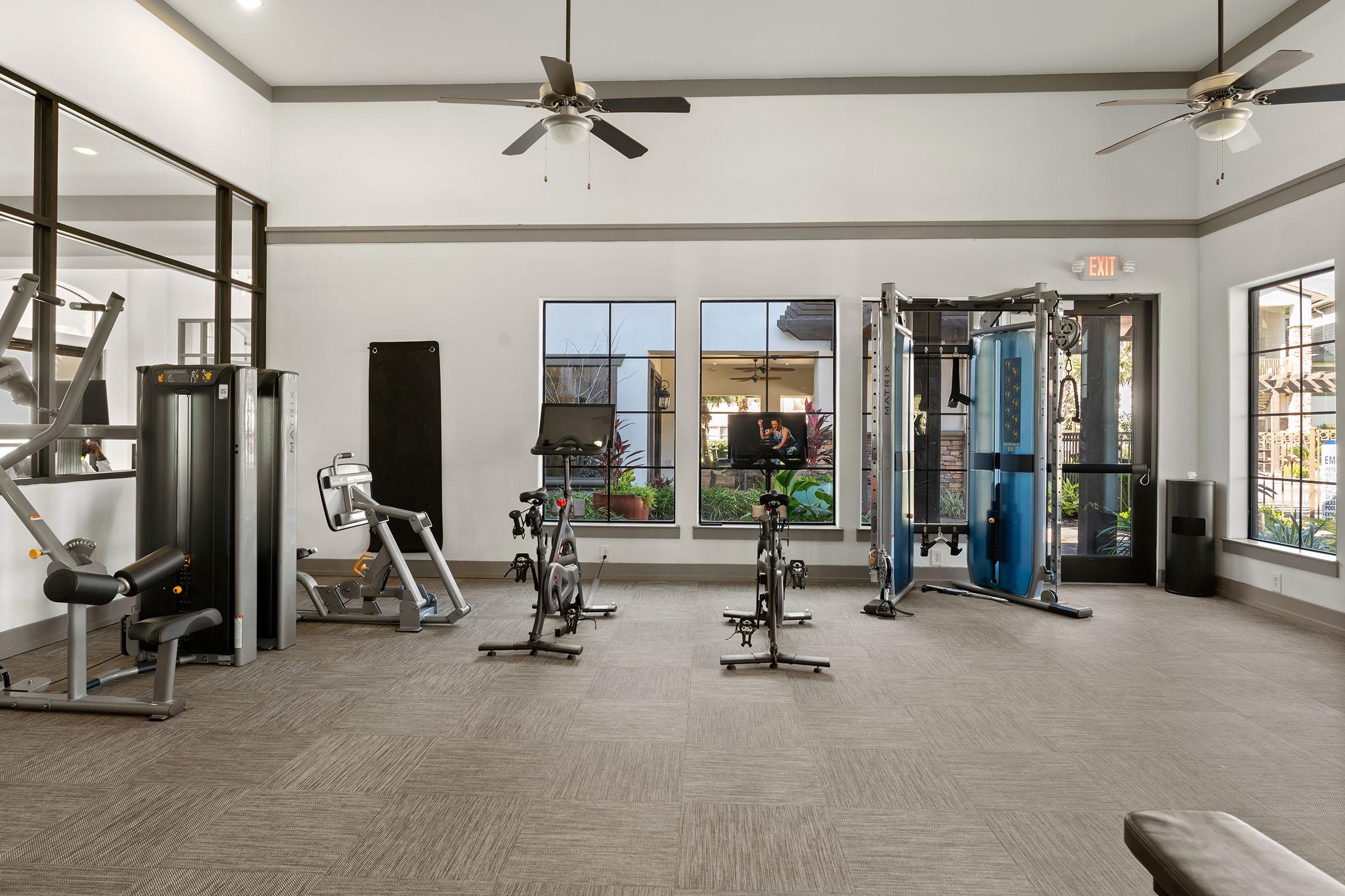 A well-lit fitness room featuring various exercise equipment. Visible items include a stationary bike, a rowing machine, a multi-use strength training machine, and a few free weights. Large windows provide natural light, and there are ceiling fans to enhance airflow. The flooring is a neutral-toned carpet.