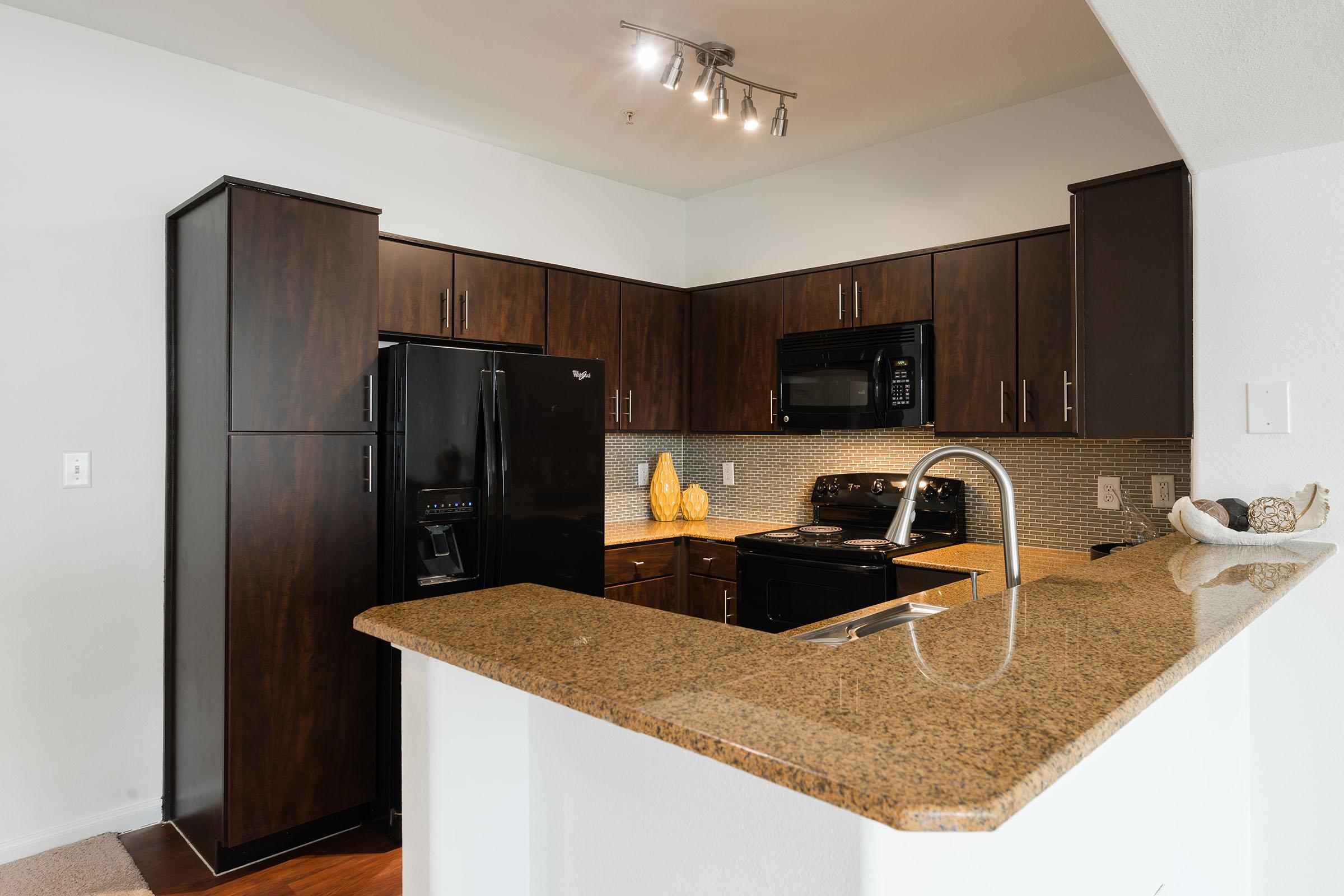 A modern kitchen featuring dark wooden cabinetry, a black refrigerator and microwave, and a granite countertop. The kitchen is well-lit with track lighting and has a mosaic tile backsplash. Decorative items are placed on the countertop, adding a touch of style to the space. The floor is a light-colored wood.