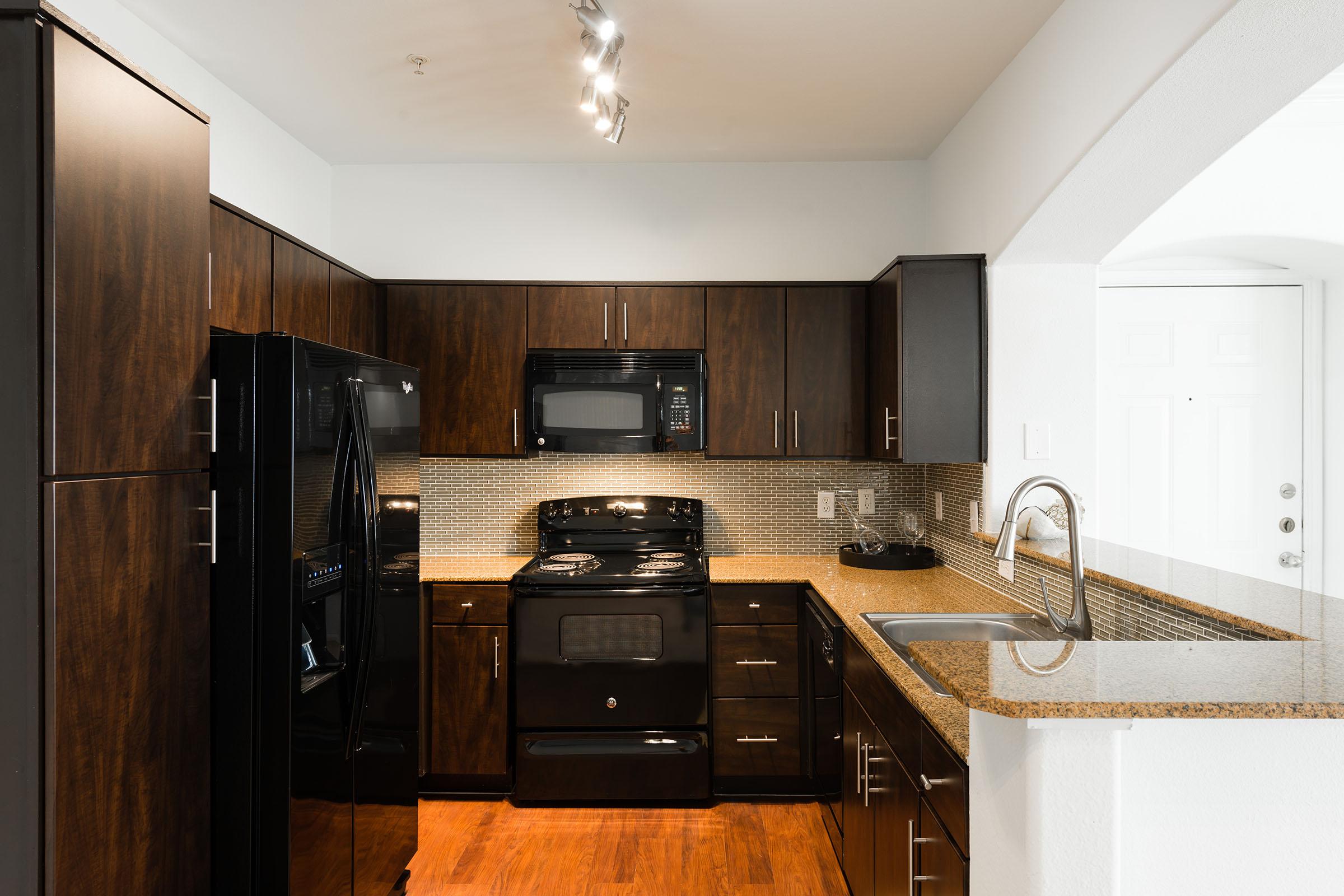 A modern kitchen featuring dark wood cabinetry and black appliances, including a refrigerator, microwave, and stove. There is a granite countertop with a stainless steel sink. The backsplash consists of small tiles, and the flooring is a warm wood tone. The kitchen is well-lit with overhead lighting.