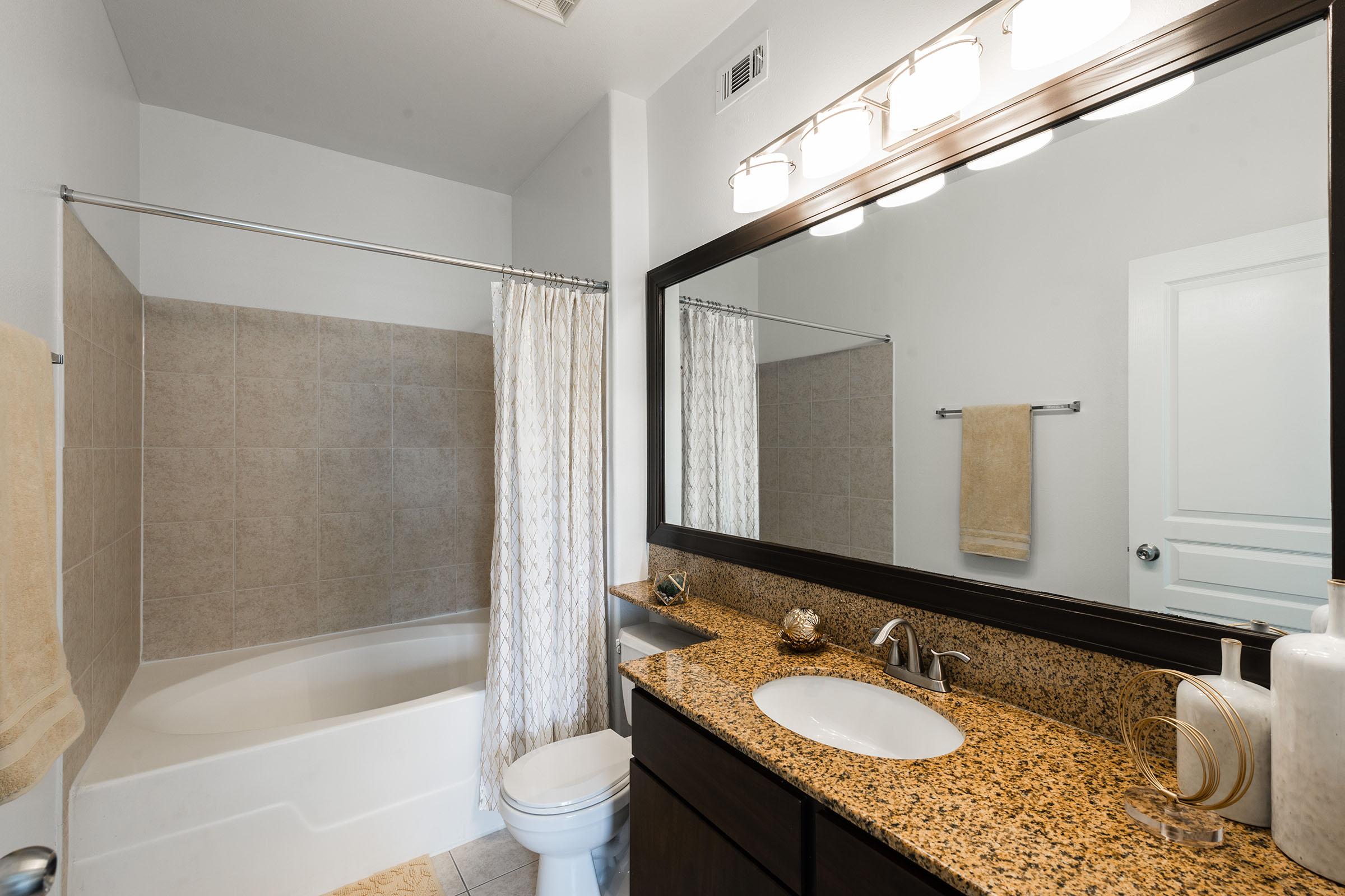 A modern bathroom featuring a bathtub with a shower curtain, a large mirror above a granite countertop sink, and a toilet. The walls are light-colored, and there are beige tiles in the shower area. A towel hangs on a rack, and decorative items are arranged on the countertop. Bright lighting is provided by three overhead fixtures.