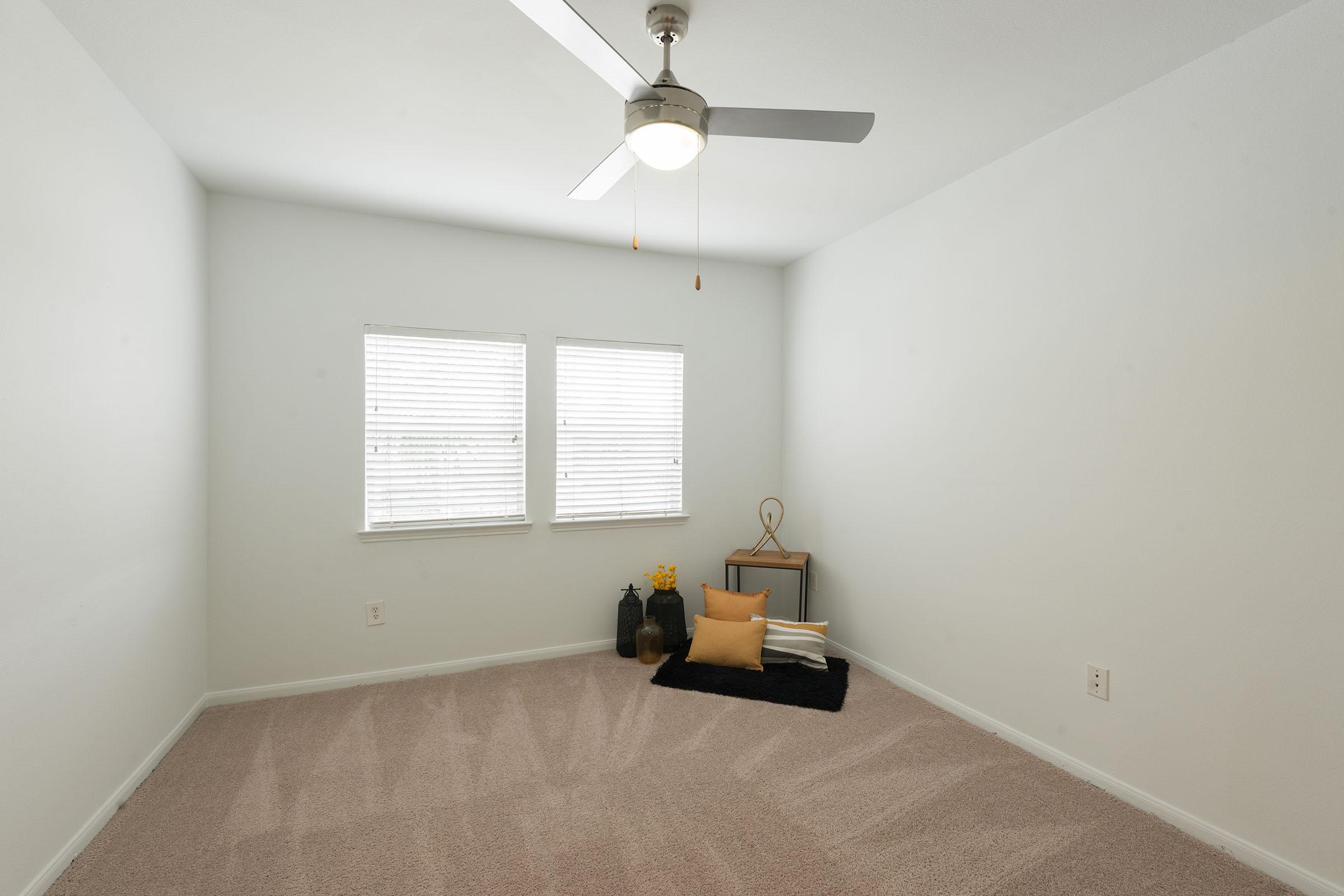 A simple, empty room with light beige carpet and white walls. There are two windows with blinds letting in natural light. A ceiling fan is visible, and in one corner, there's a small decorative arrangement featuring a black mat, a vase, yellow flowers, and a throw pillow.