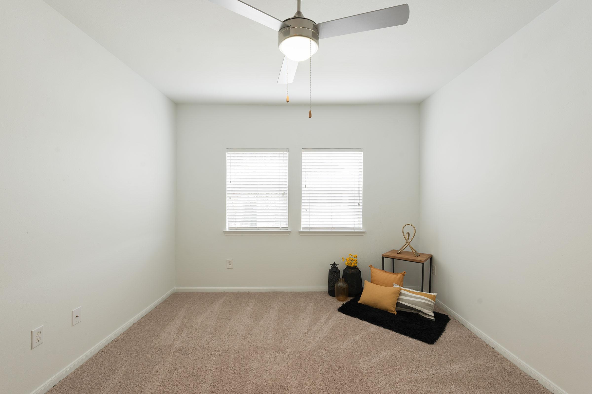 A minimalistic room featuring beige carpet, two windows with white blinds, and a ceiling fan. In one corner, a small table with decorative items and a black rug with yellow cushions. The walls are painted in a light color, creating an open and airy atmosphere.