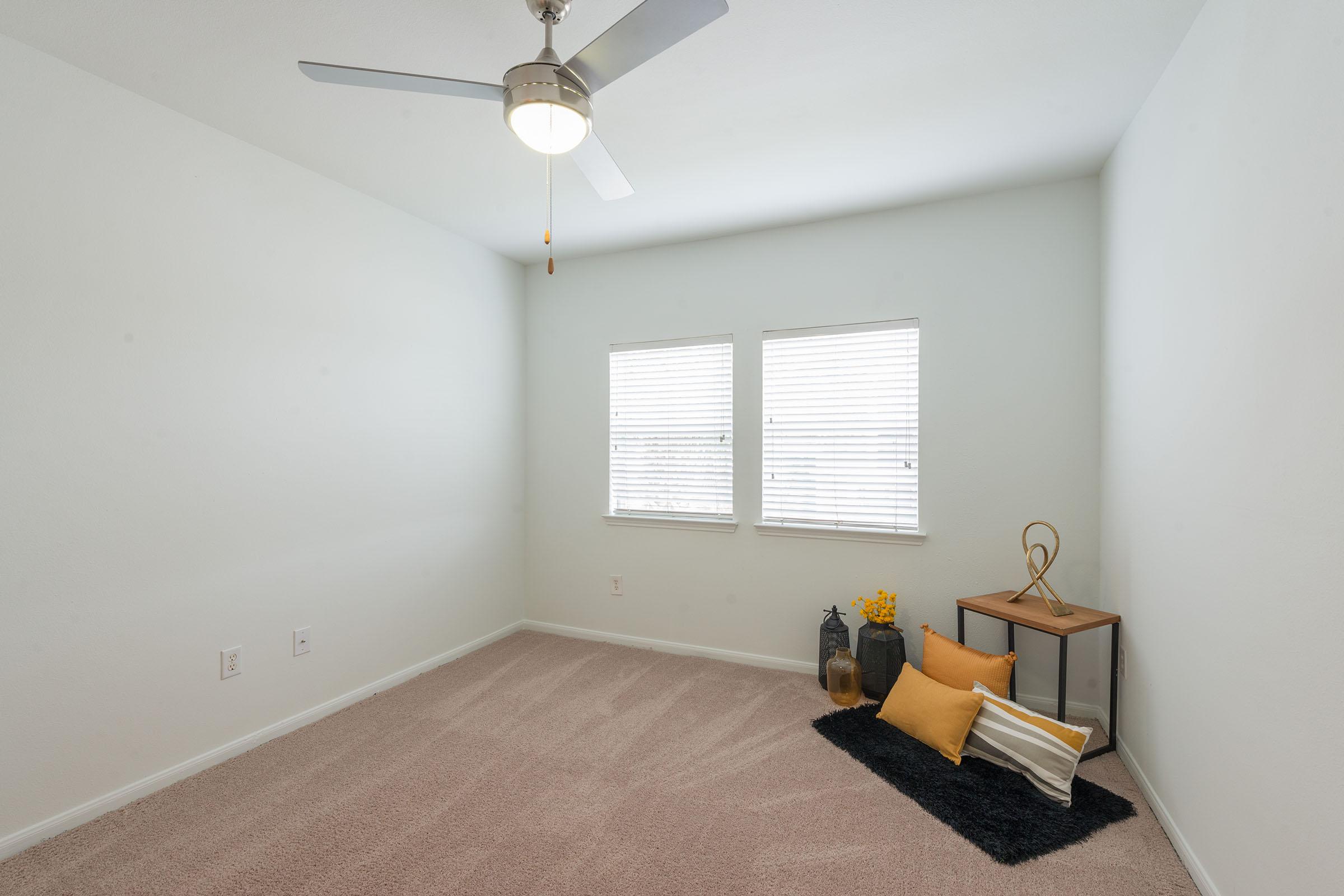 Empty room with light-colored walls and carpet, featuring two windows with white blinds. A ceiling fan is installed. There is a small wooden table with decorative items, including a vase and two jars, and a black rug with a yellow pillow and a throw blanket on the floor.