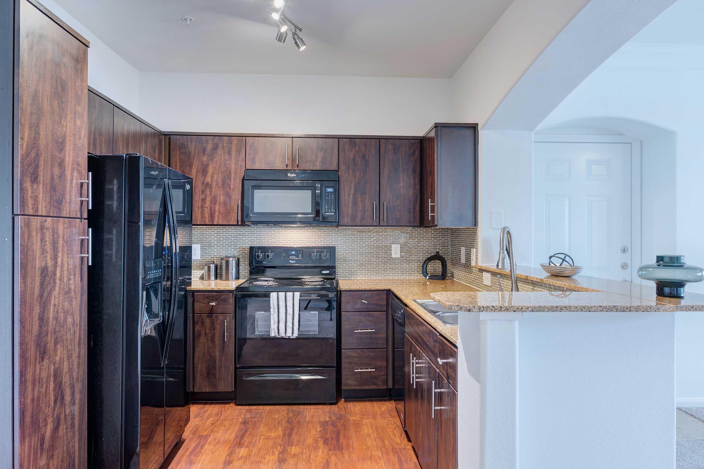 Modern kitchen featuring dark wood cabinetry, a black refrigerator, and a black stove with microwave. The counter is made of light granite, and there is a neutral-colored backsplash. The floor is wooden, and a doorway is visible in the background, suggesting an open layout.