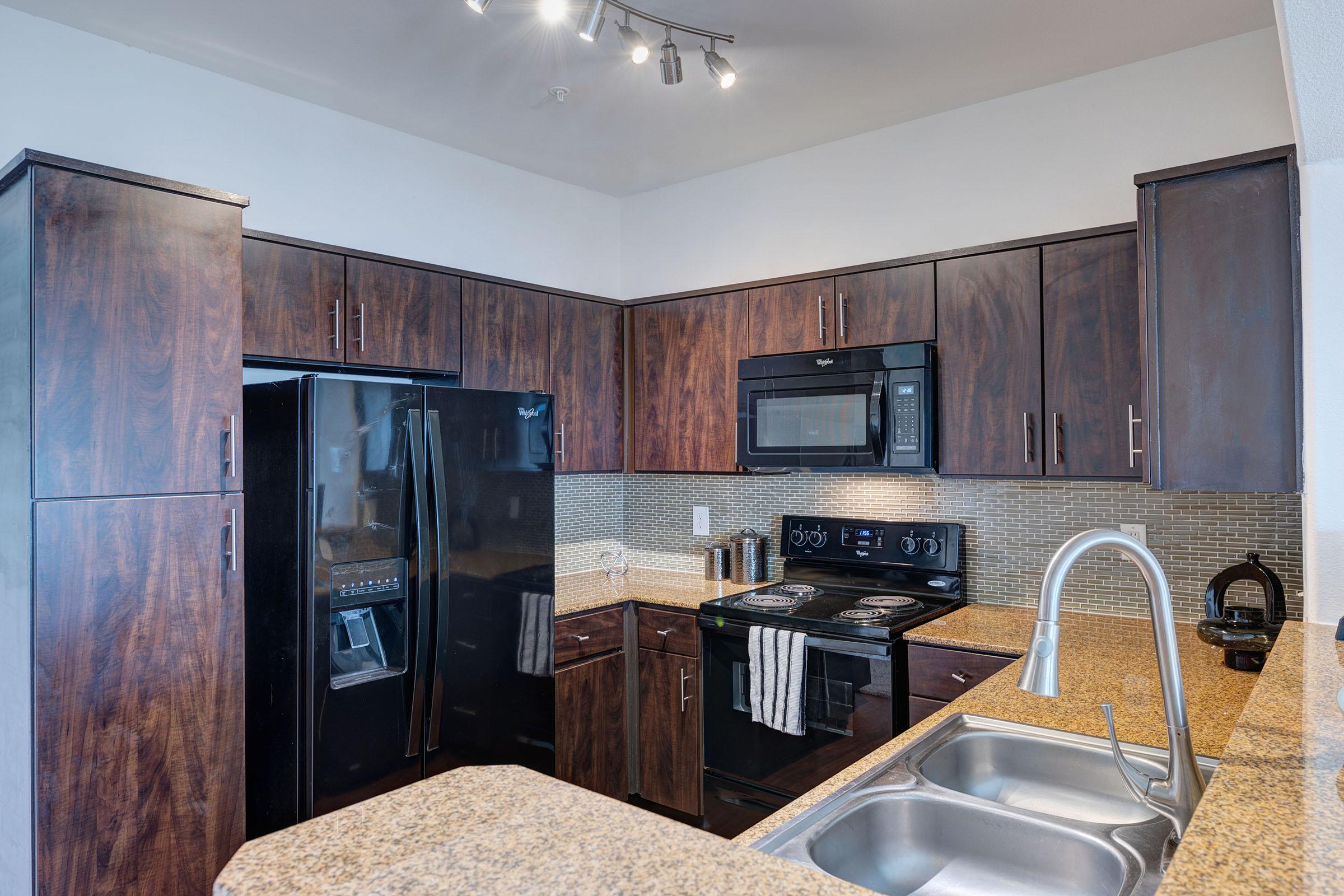 Modern kitchen featuring dark wood cabinets, black appliances including a refrigerator and microwave, granite countertops, and a sink with a brushed nickel faucet. The kitchen is well-lit with overhead track lighting and includes a mosaic tile backsplash.