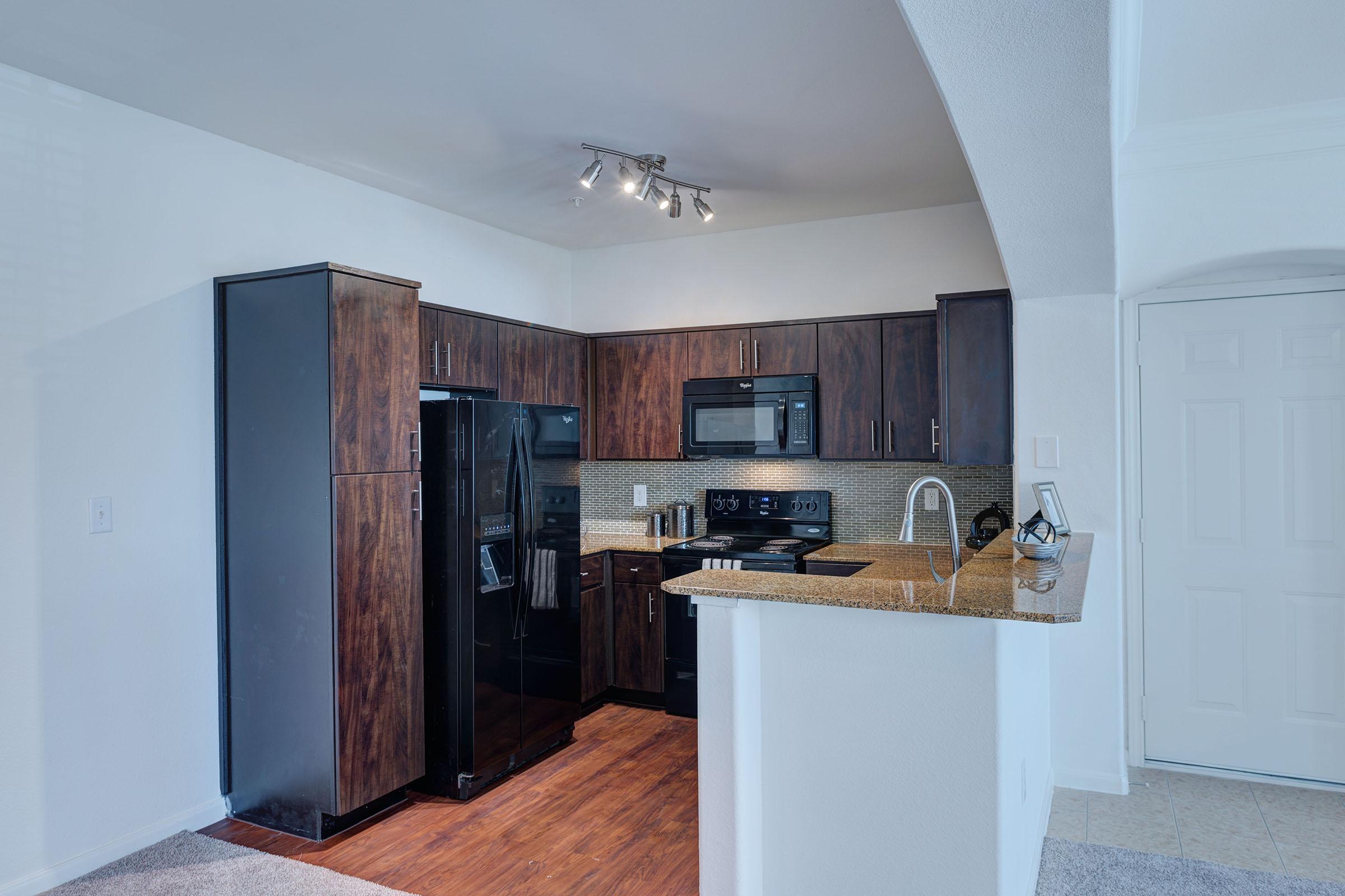 Modern kitchen featuring dark wooden cabinets, black appliances, and granite countertops. The space includes a built-in microwave, oven, and stovetop, with track lighting above. There’s a breakfast bar open to the living area, and neutral-colored walls and flooring enhance the contemporary feel.