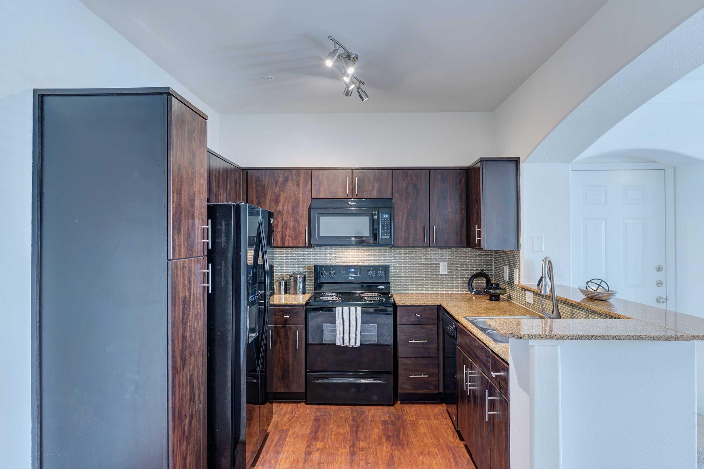 Modern kitchen featuring dark wood cabinetry, a black refrigerator, and an integrated microwave above the stove. The countertops are light-colored granite, and the space is well-lit with track lighting. A sleek design complements the hardwood floor and a partial archway leads to another room.