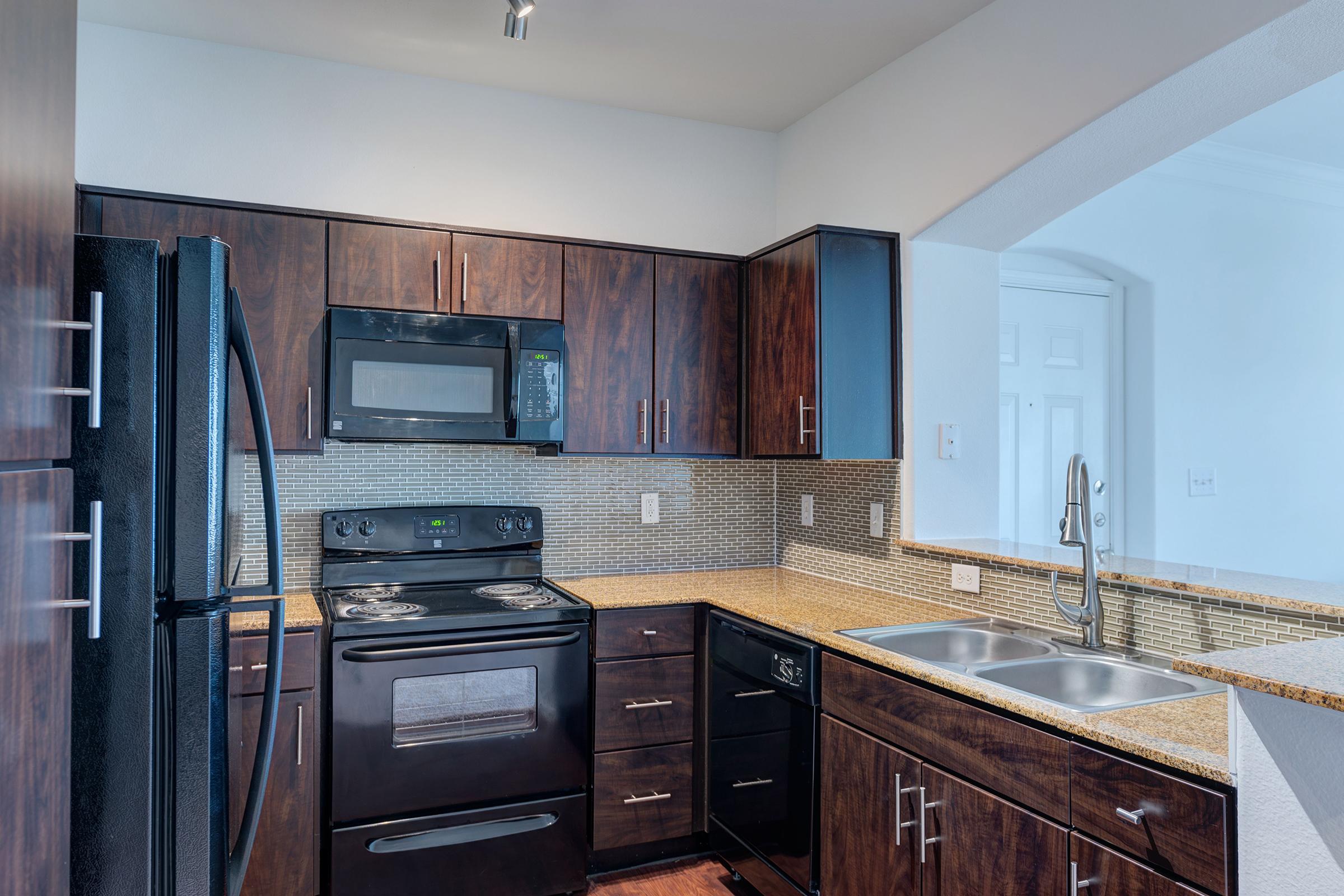 Modern kitchen featuring dark wood cabinetry, a black refrigerator, stove, and microwave, with stainless steel accents. The countertops are light granite, and there are dual sinks. A decorative tile backsplash adds a stylish touch, and an arched entryway leads to another room.