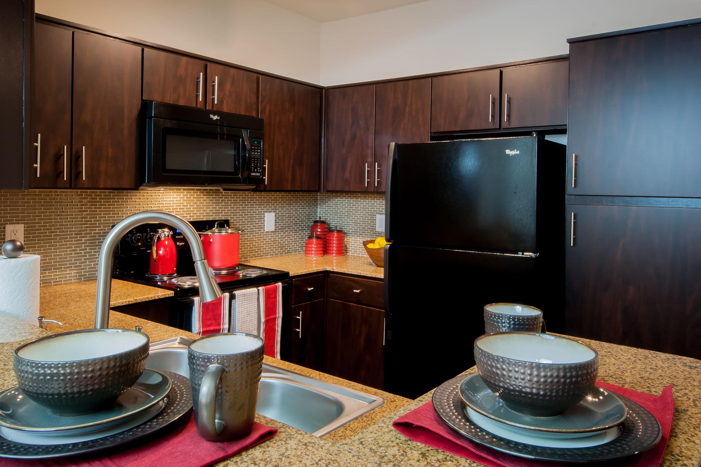 Modern kitchen featuring dark wooden cabinets, a black refrigerator, and a black microwave. The countertop is made of light granite, with a sink in the foreground. Two plates and mugs are set on red napkins, and a bowl of fruit is visible next to a red kettle on the stove. Subtle tile backsplash adds texture to the space.