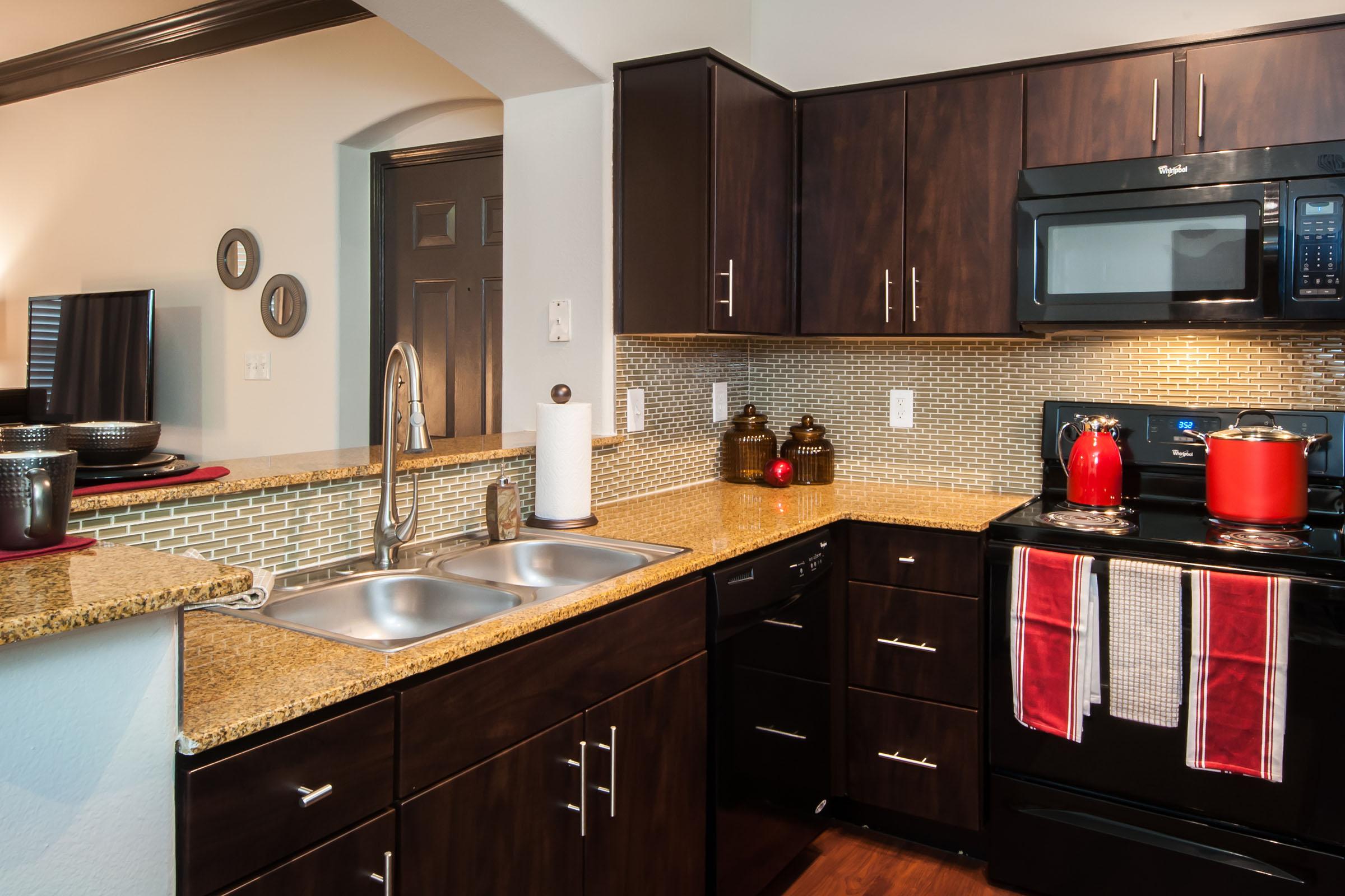 A modern kitchen featuring dark wooden cabinets, a granite countertop with a double sink, a black oven and microwave, and red accents. The backsplash is made of small tiles, and there are decorative items, including a paper towel holder and two red pots on the stove. Natural light from a nearby window enhances the space.