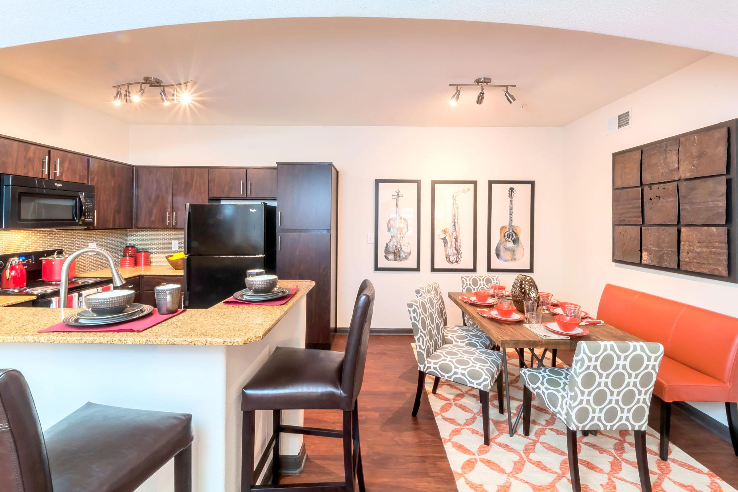 Modern kitchen and dining area featuring a granite countertop, dark wooden cabinets, and a black refrigerator. The dining table is set with red and white dishes, surrounded by stylish chairs. Wall art includes framed guitars and abstract pieces. Warm lighting enhances the inviting atmosphere.