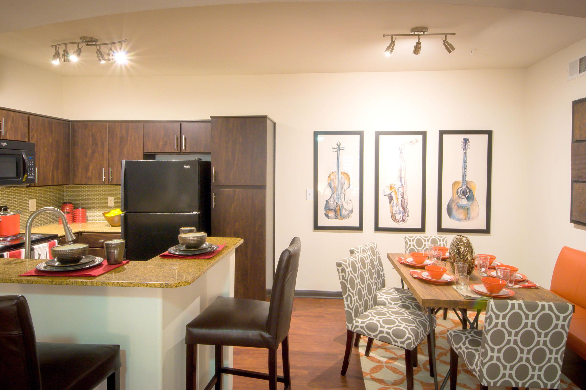 A modern kitchen and dining area featuring dark wood cabinets, a black refrigerator, and sleek countertops. The dining table is set with red bowls and decorated with a centerpiece, surrounded by patterned chairs. Three framed musical instrument artworks adorn the wall, adding character to the space.