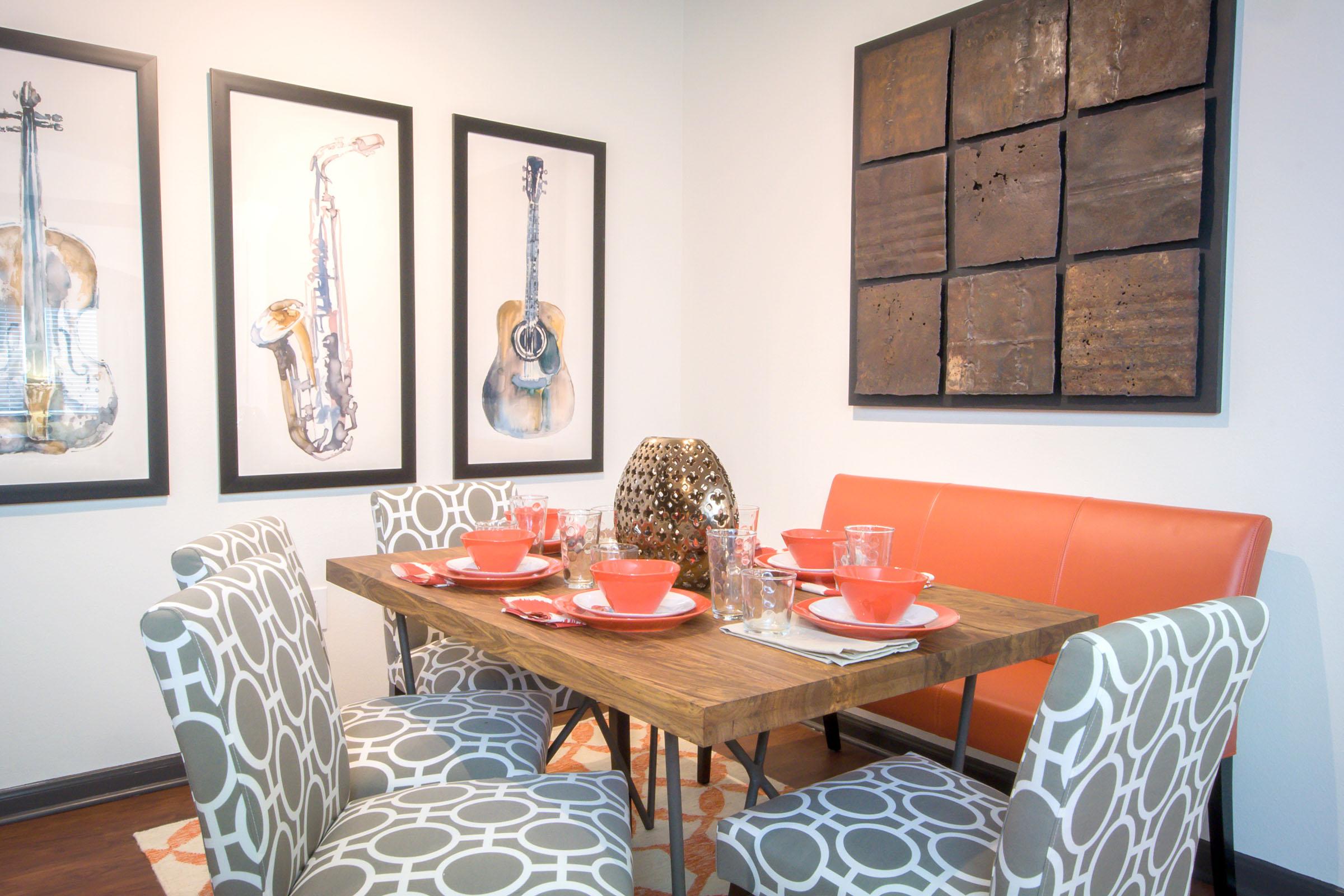 A dining area featuring a wooden table set with orange bowls, glasses, and utensils. Surrounding the table are patterned gray chairs. On the walls, there are three framed watercolor paintings of musical instruments and a large abstract artwork. The space is bright and modern.