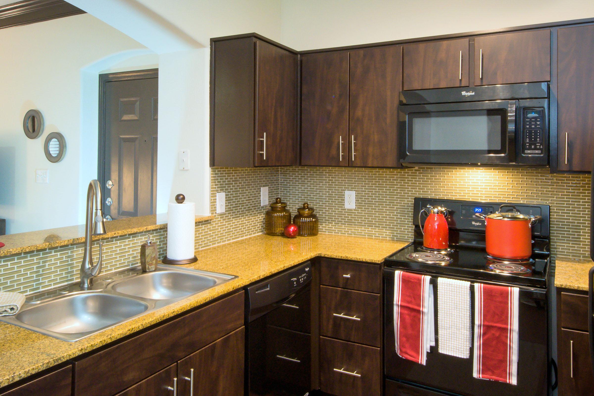 A modern kitchen featuring dark wood cabinetry and a granite countertop. The sink has two basins, and there's a gray microwave above a black stove. Red pots and towels add a pop of color, while the backsplash consists of small mosaic tiles. A door and round wall mirror are visible in the background.