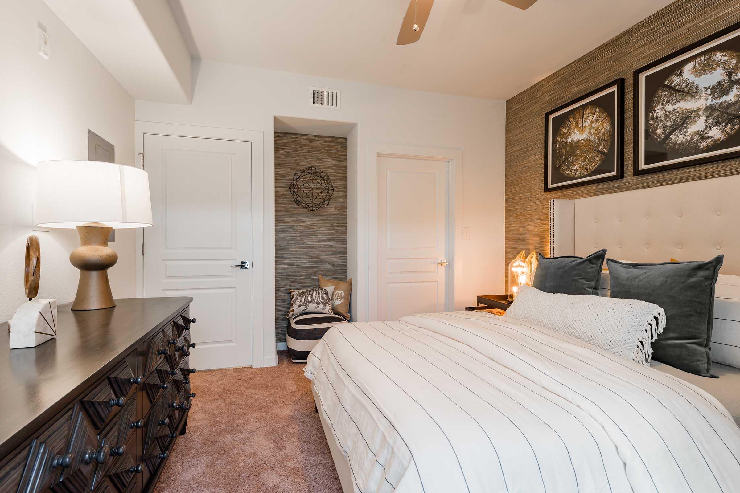 A cozy bedroom featuring a light-colored bed with striped bedding, a stylish headboard, and a dresser. The walls are adorned with a textured wallpaper and framed artwork. A ceiling fan is visible, along with a small decorative table lamp. There's a door leading to another room and a cozy nook with pillows.